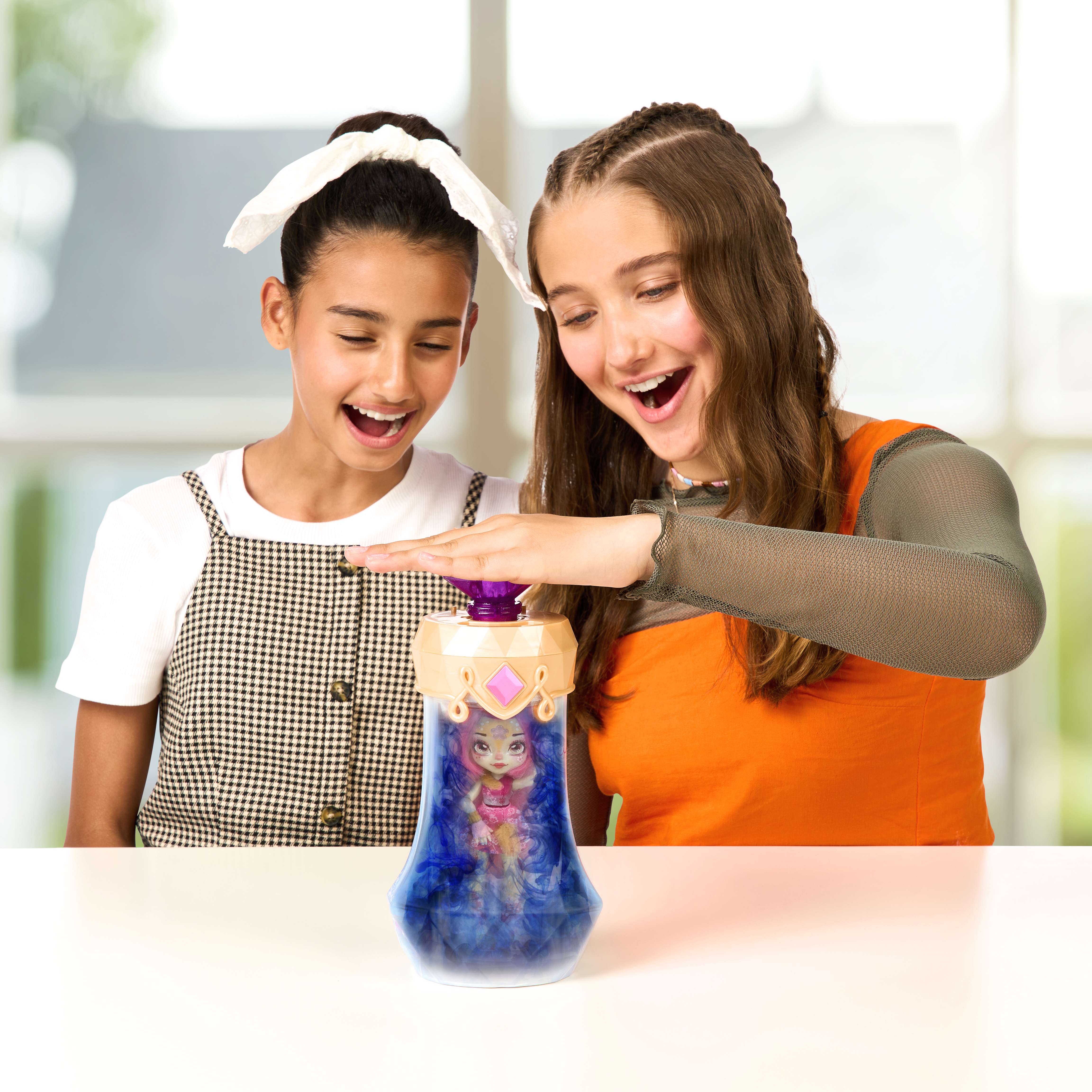 Dwie dziewczyny patrzą na zabawkę. Jedna dziewczyna naciska pokrywkę zabawki. Zabawka stoi na białym stole.