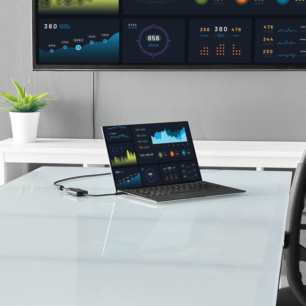 Laptop wyświetla wykresy danych, połączony z ekranem pokazującym metryki. Roślina w białej doniczce stoi na stole.
