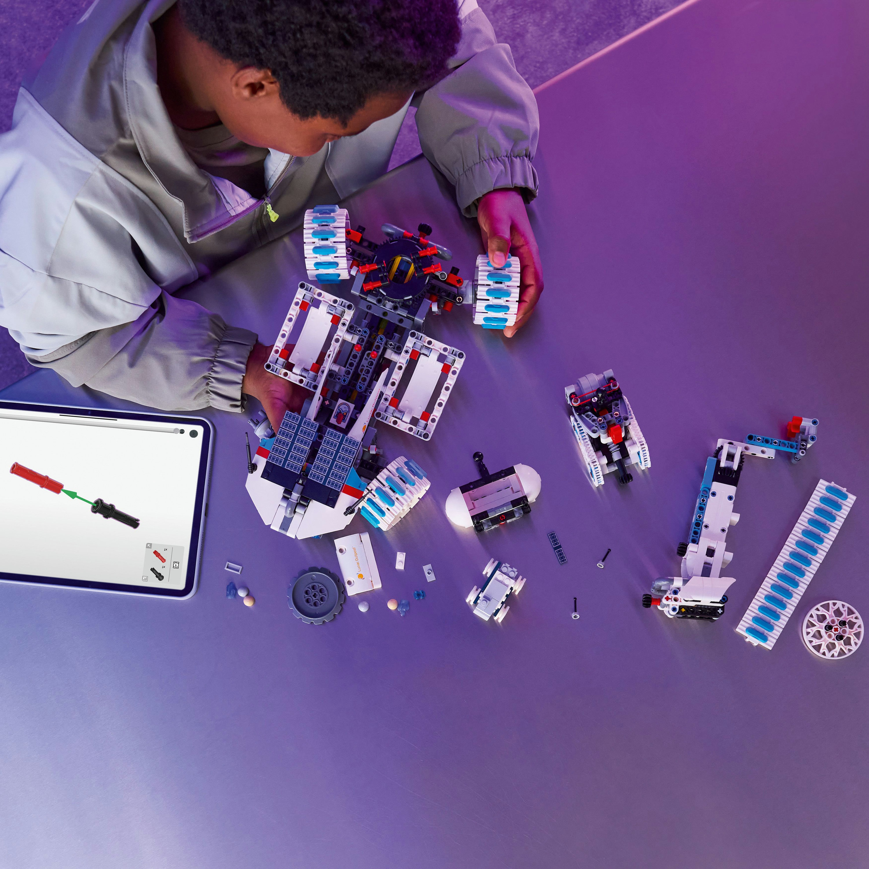 Osoba montuje model Lego Technic. Scena obejmuje części, tablet z instrukcjami i fioletową powierzchnię.