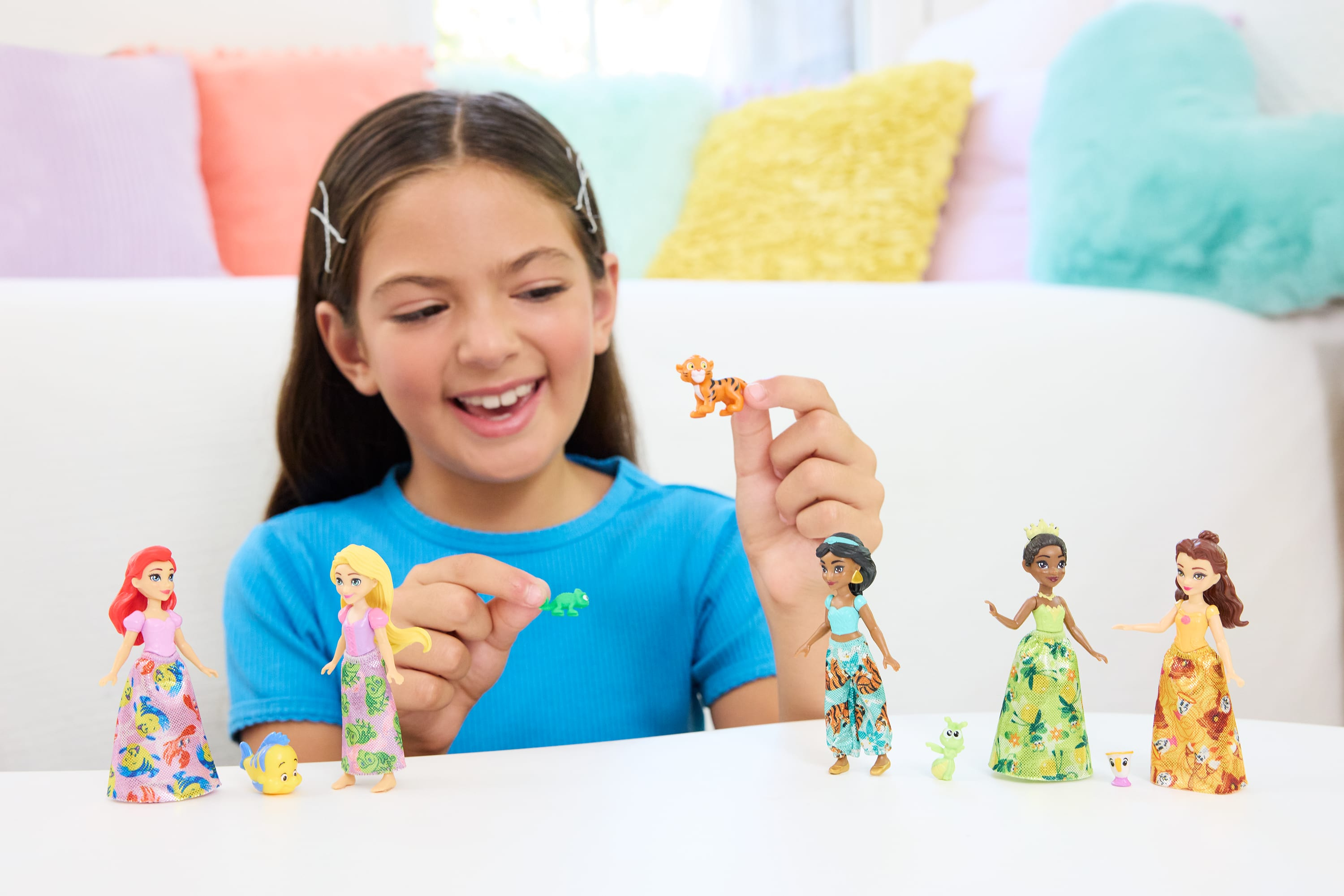 Dziewczyna uśmiecha się, trzymając figurki. Lalki księżniczek i zabawka tygrysa na białym stole. Poduszki w tle.