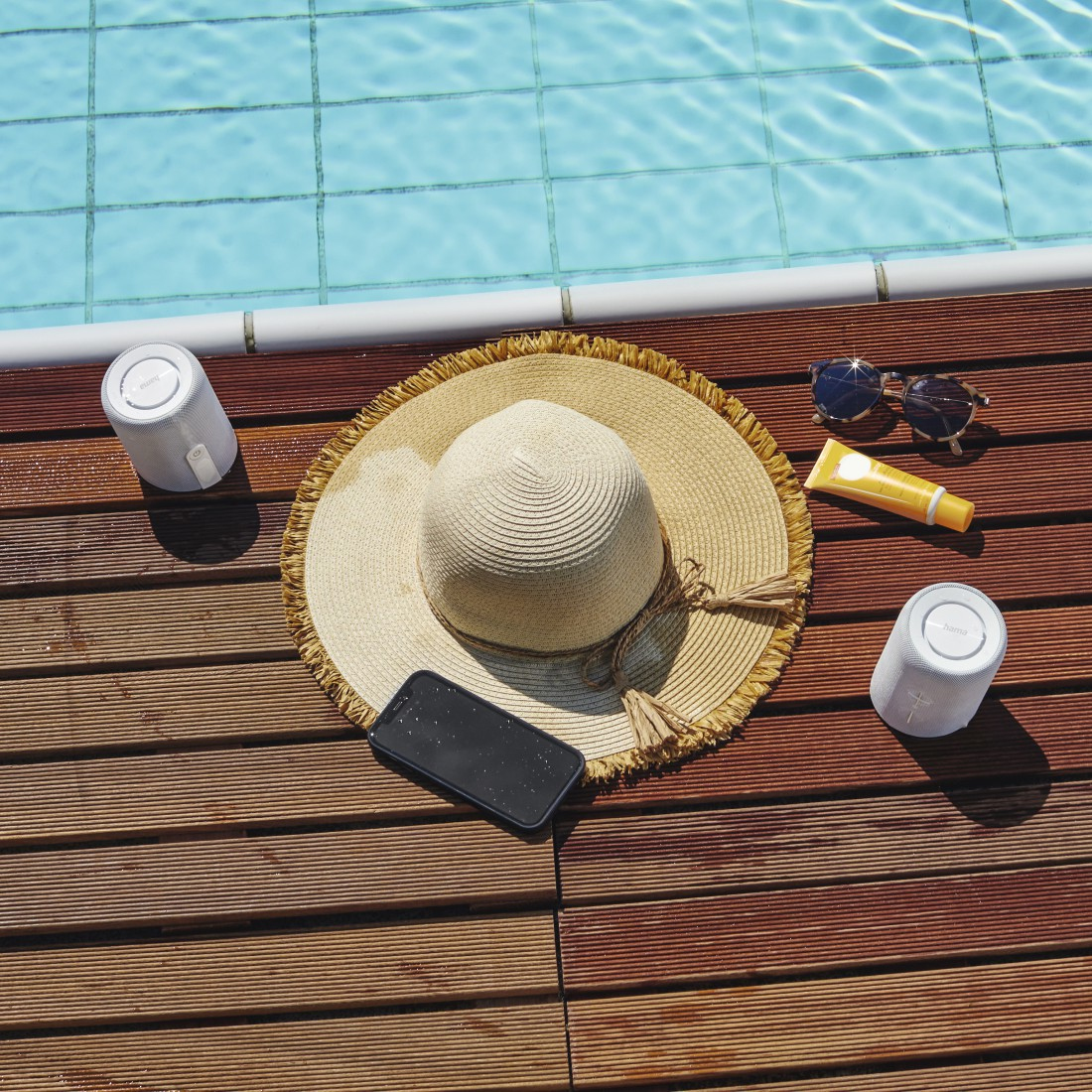 Kapelusz przeciwsłoneczny, głośniki, telefon, okulary przeciwsłoneczne i krem ​​do opalania leżą na drewnianym pokładzie przy basenie.