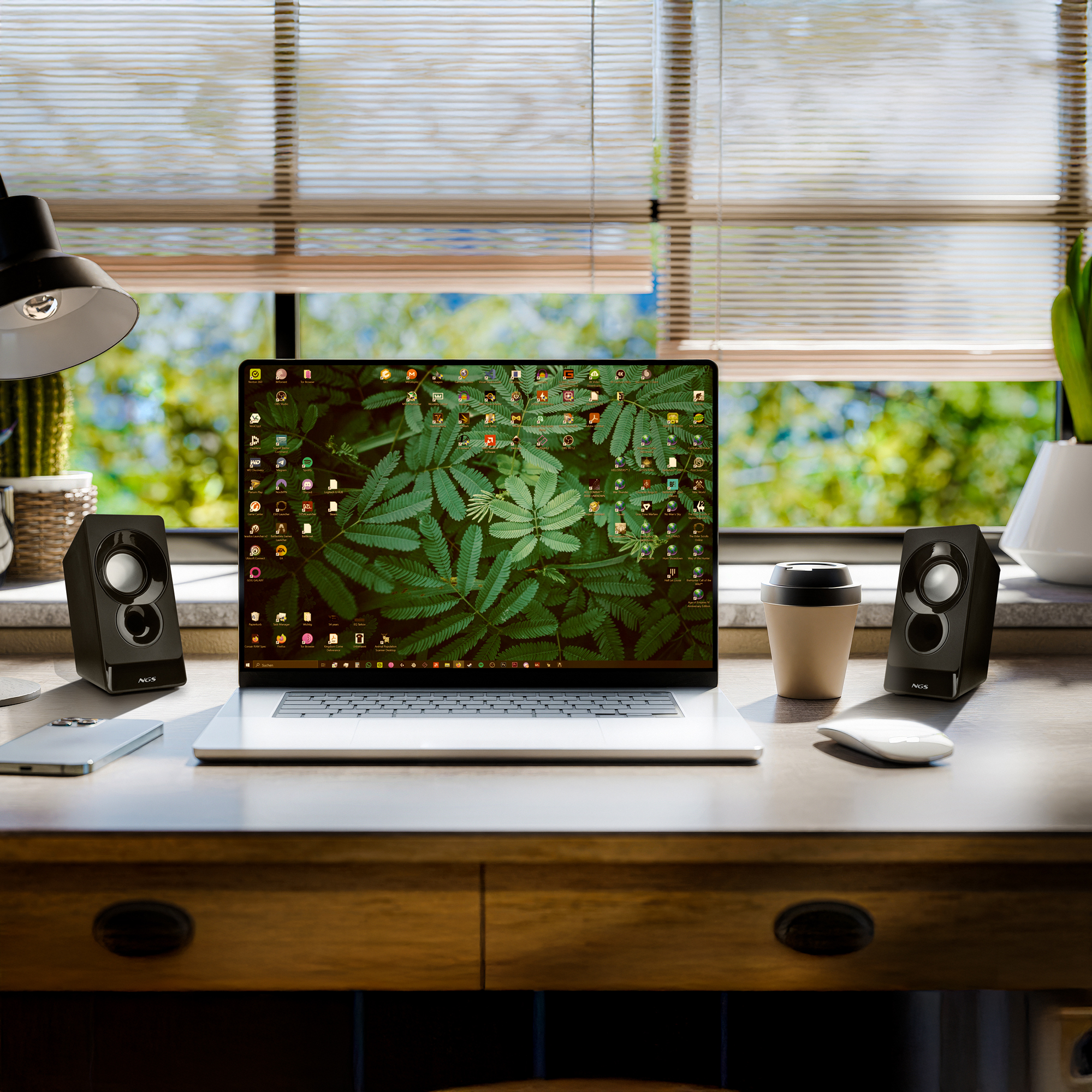 Laptop auf dem Schreibtisch mit Lautsprechern, Kaffeetasse und Maus. Jalousien und ein Fenster mit grünem Laub im Hintergrund.