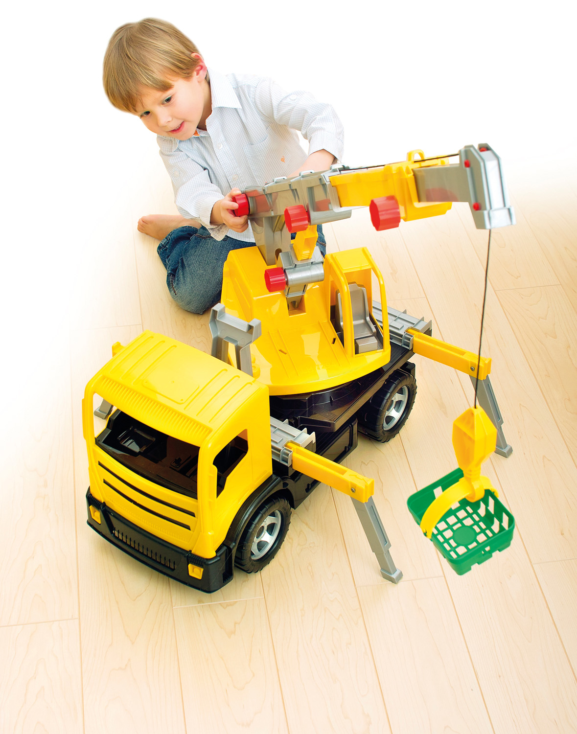 Un bambino gioca con un camion gru giocattolo giallo. La gru ha un cestino verde. È su un pavimento di legno, con sfondo bianco.