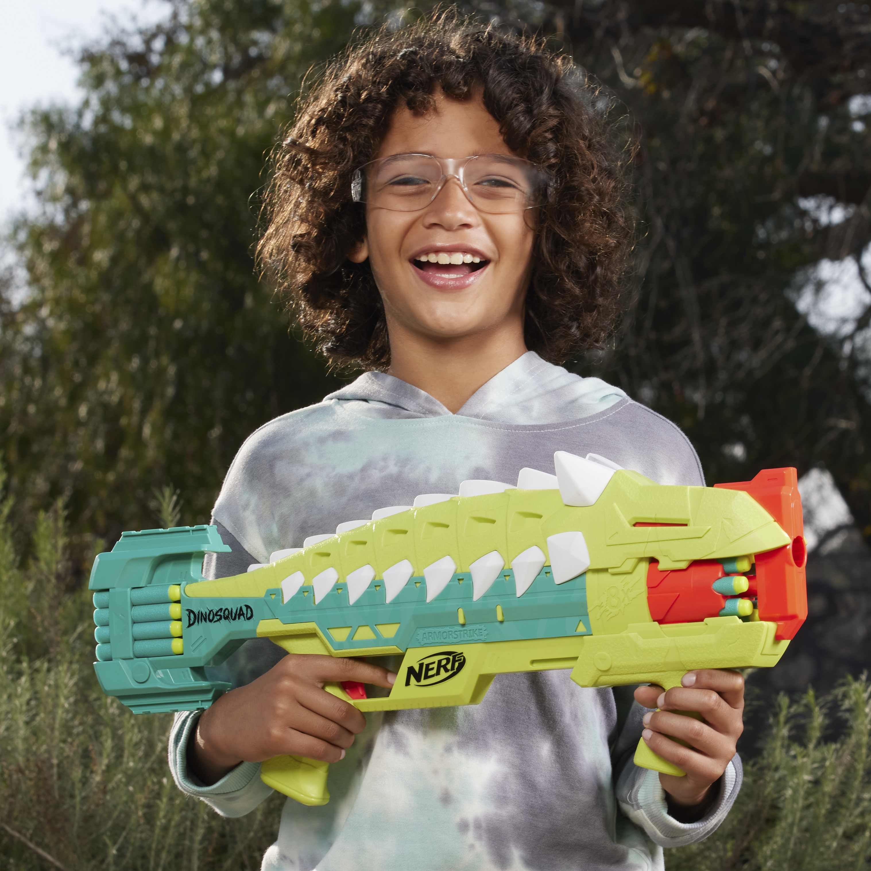 Un niño sonriente sostiene una pistola Nerf verde y amarilla con temática de dinosaurios. Lleva gafas y una sudadera gris, con follaje de fondo.