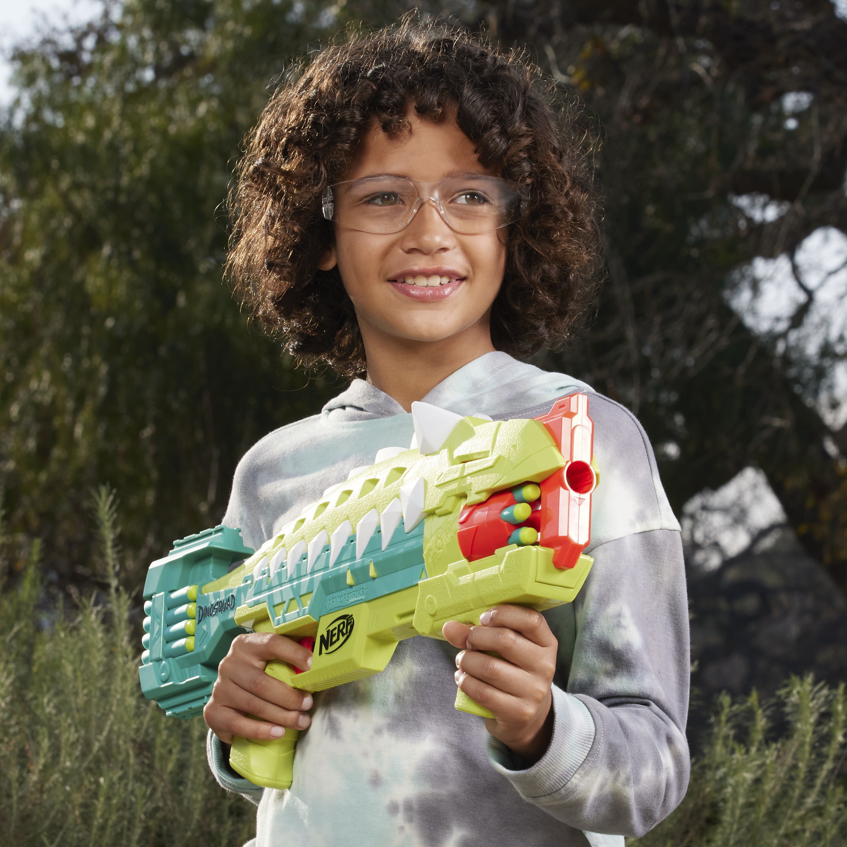Un niño sonríe, sosteniendo una pistola de juguete verde y amarilla con gafas de seguridad. El fondo son árboles verdes borrosos.