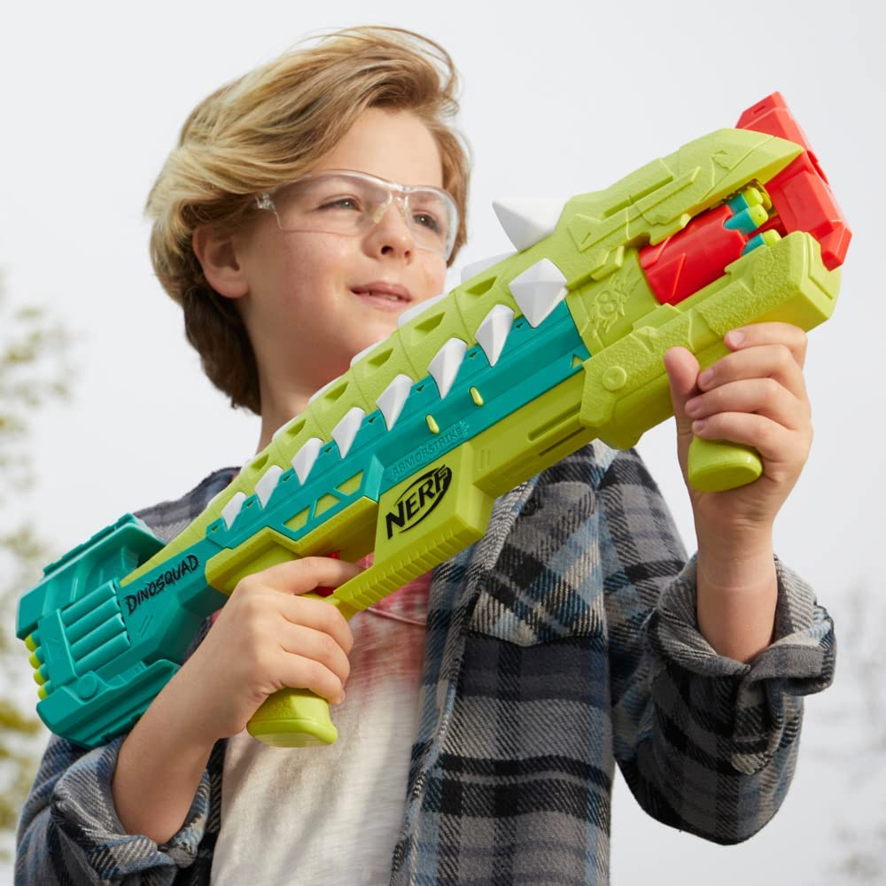 Un niño con gafas sostiene una pistola Nerf verde y azul. El arma tiene detalles blancos en forma de dientes y detalles rojos.