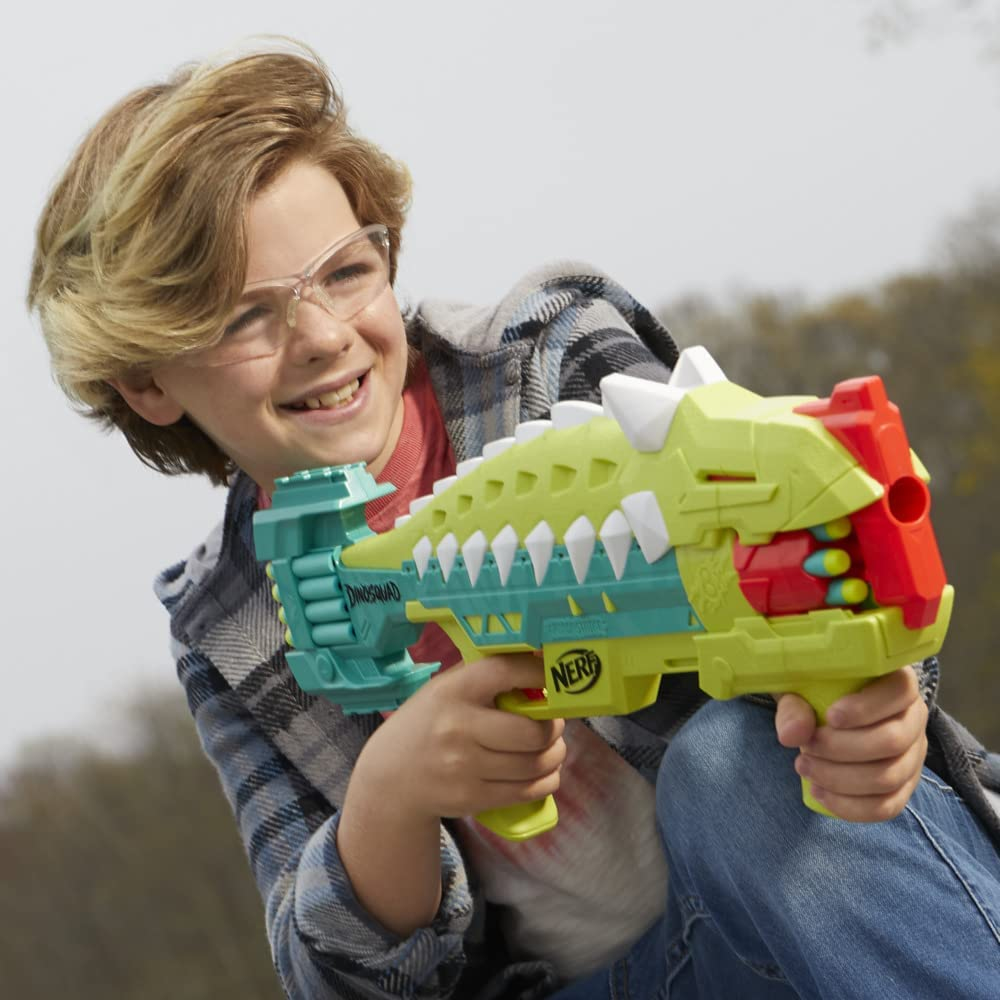 Un niño con gafas sonríe, sosteniendo una pistola Nerf con temática de dinosaurio verde y amarillo. Lleva una camisa a cuadros y vaqueros.