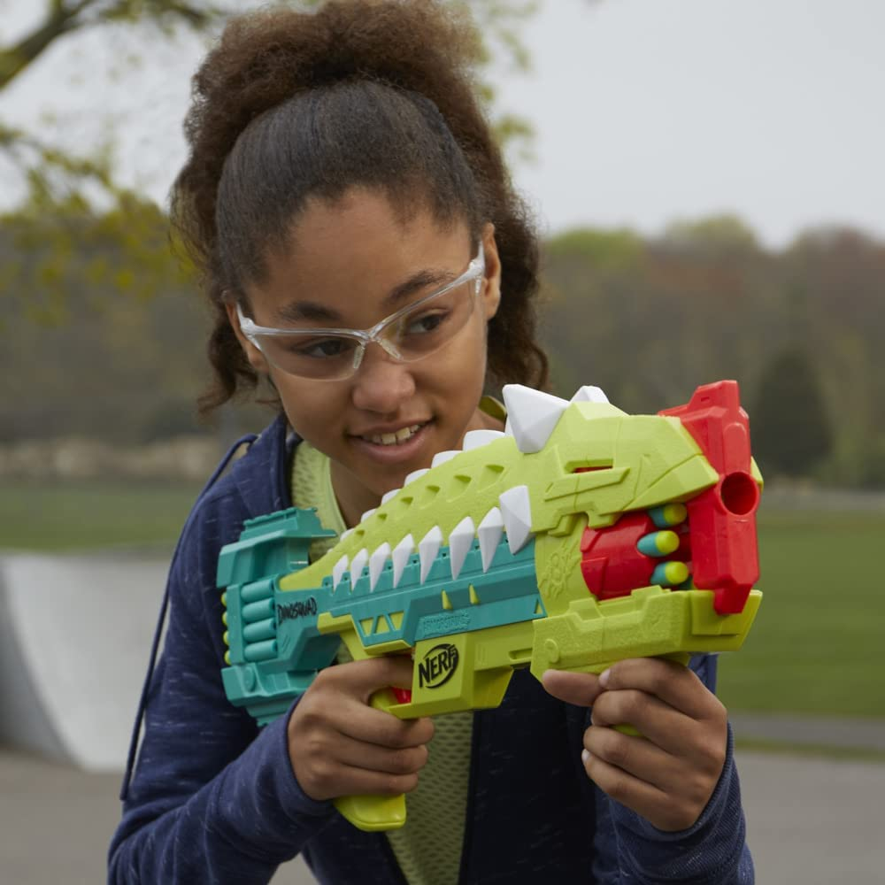 Una niña con gafas sostiene una pistola de juguete verde y roja. La pistola tiene púas blancas. Ella sonríe.