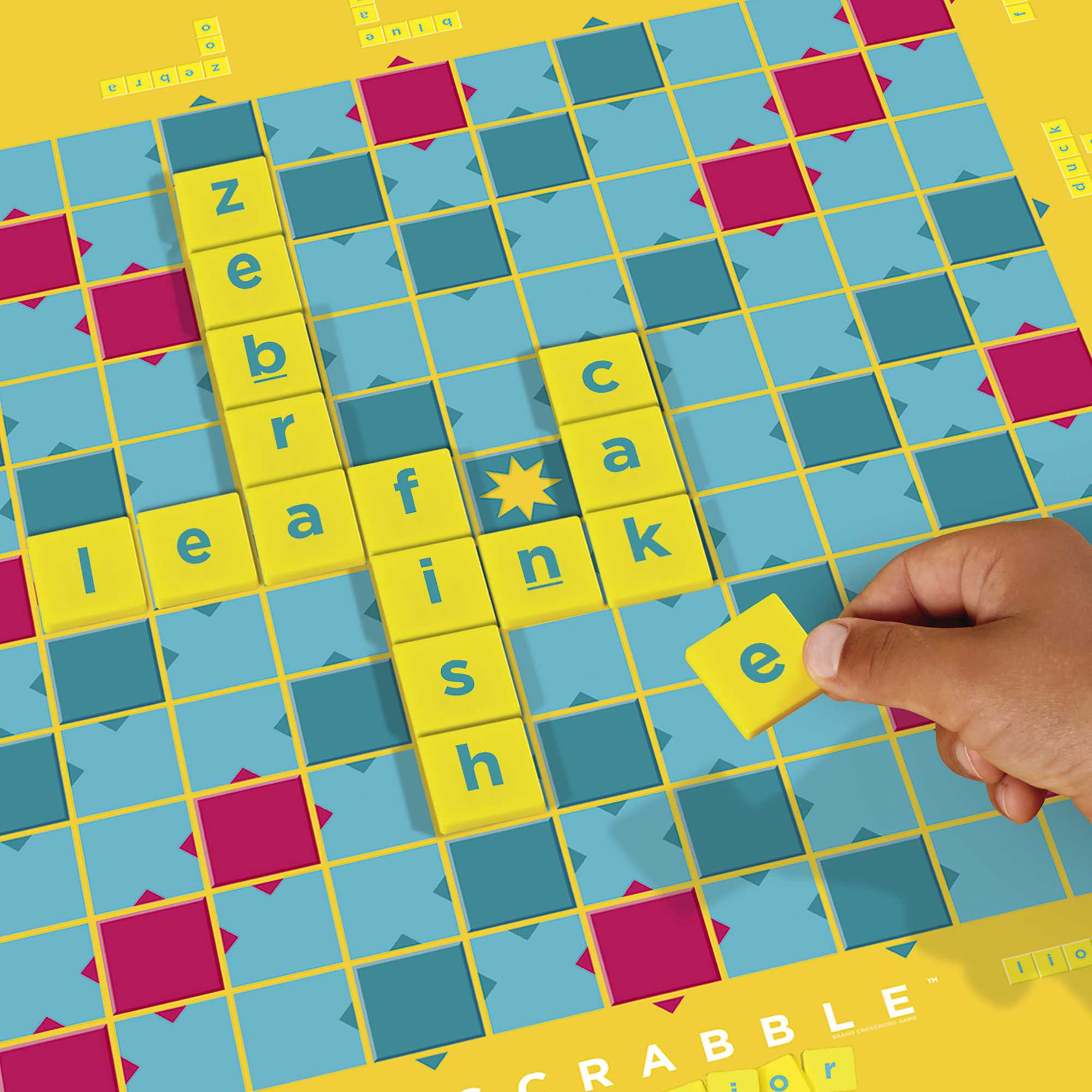 Ręka umieszczająca kafel 'e' na planszy do Scrabble. Plansza ma niebieskie, różowe i żółte kwadraty. Tworzone są słowa.