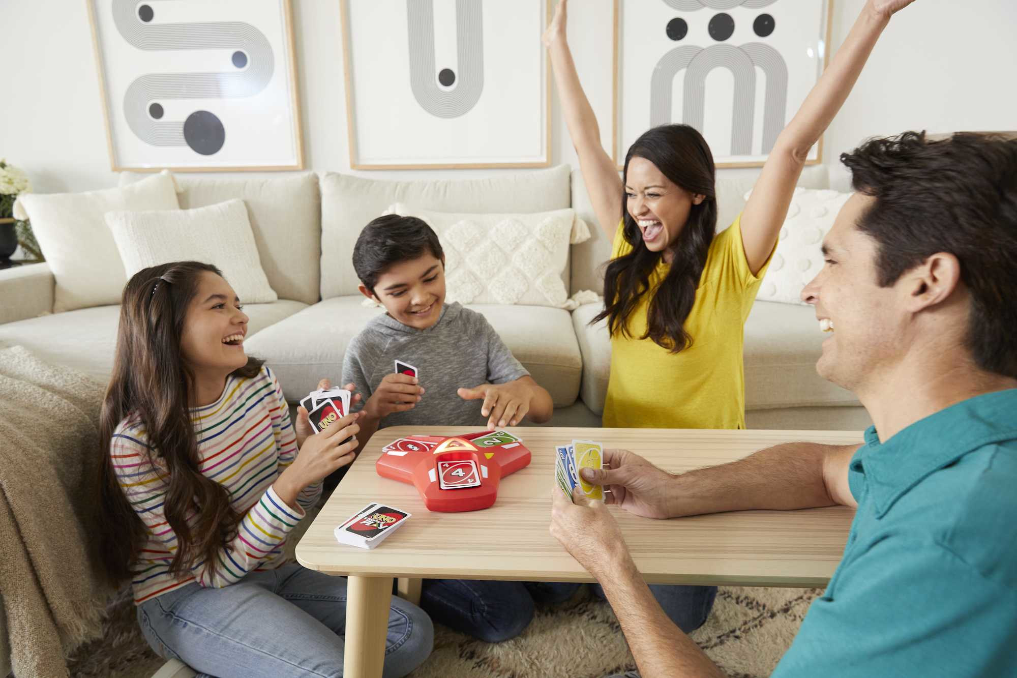 Familie spielt Uno. Eine Frau hebt die Arme zum Sieg. Andere lächeln, halten Karten und sitzen um einen Tisch.