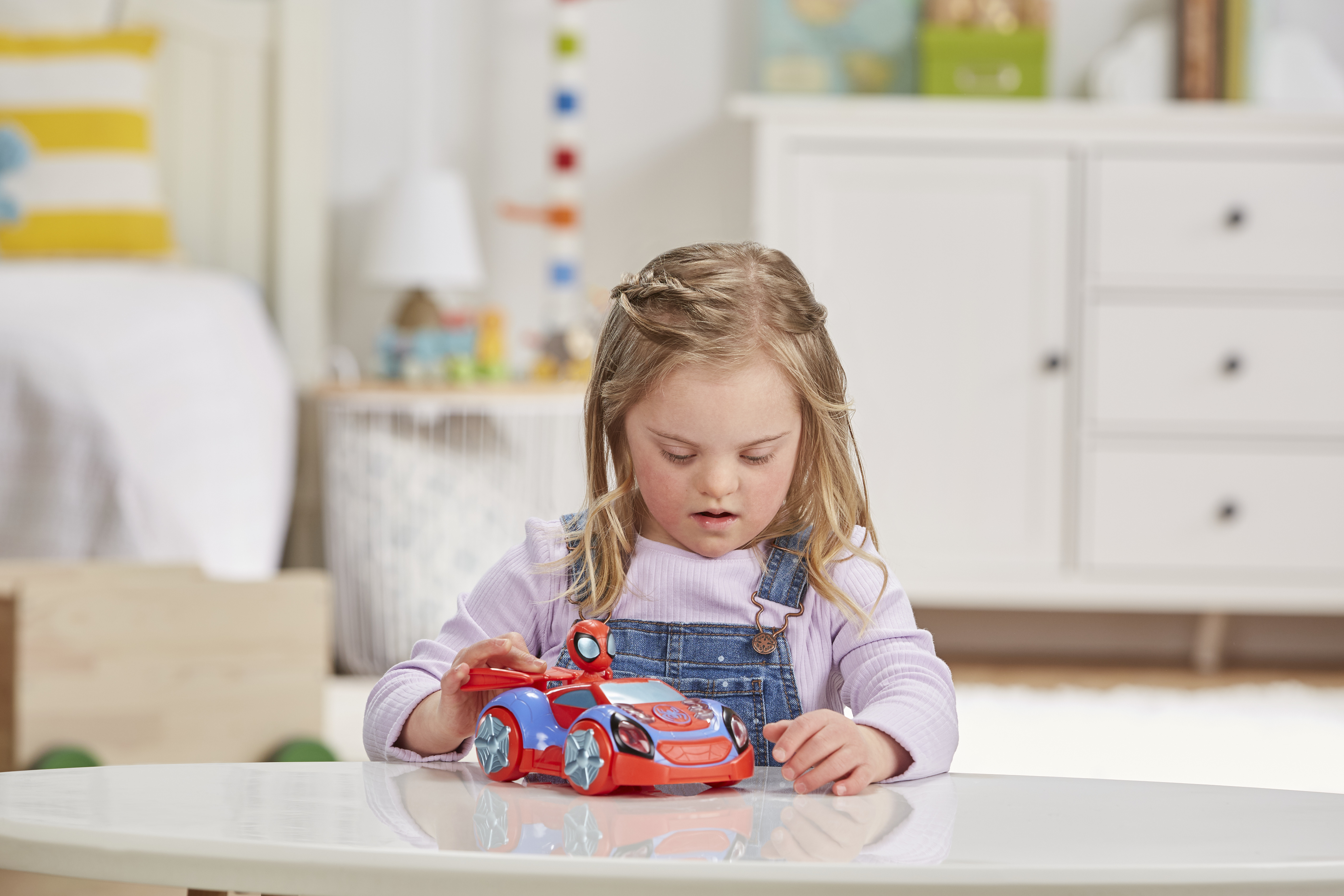 Dziewczynka z zespołem Downa bawi się samochodzikiem. Samochód jest czerwono-niebieski, a dziewczynka siedzi przy białym stole.
