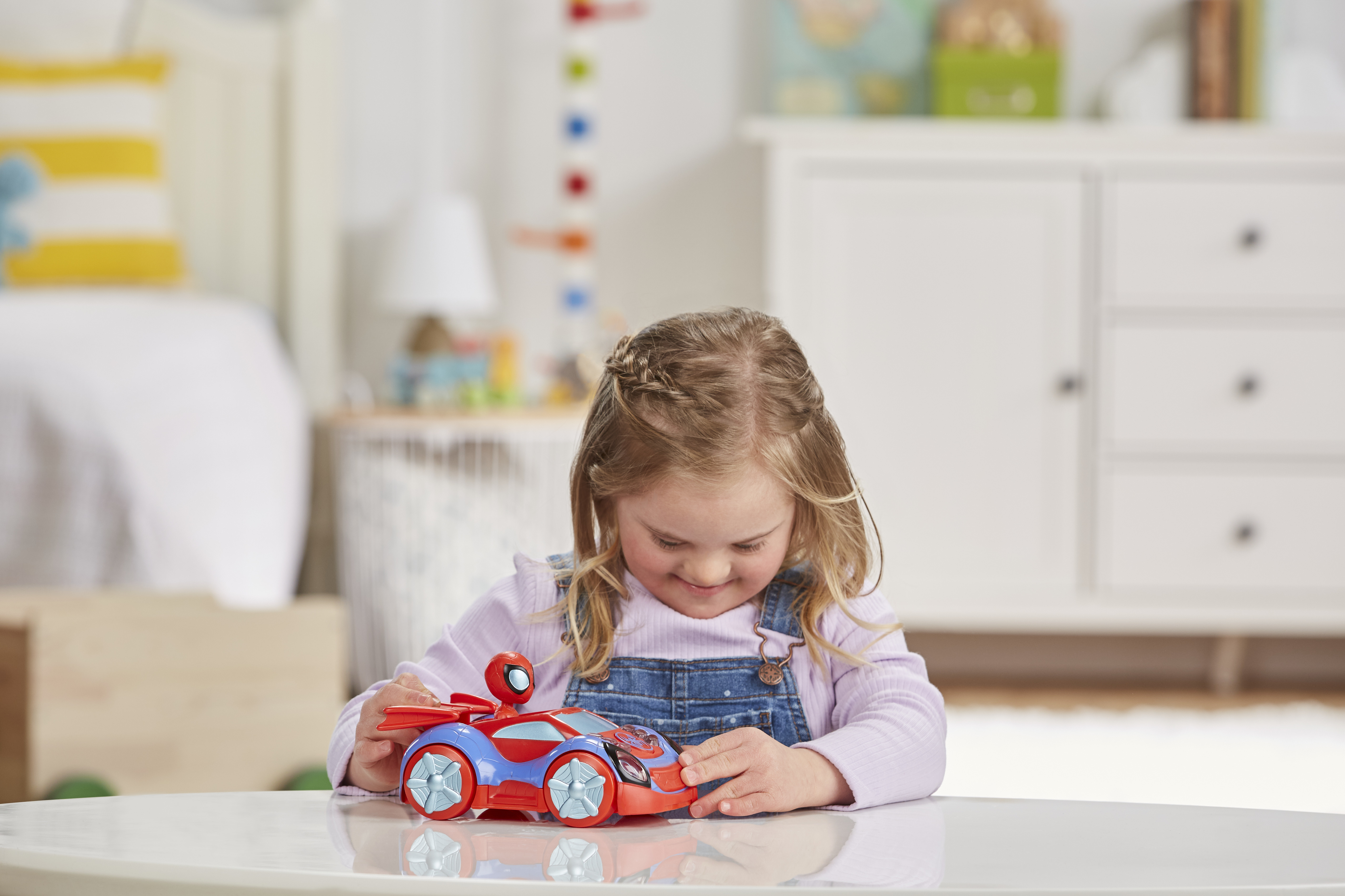 Dziewczynka z blond warkoczami uśmiecha się do czerwono-niebieskiego samochodziku na białym stole.