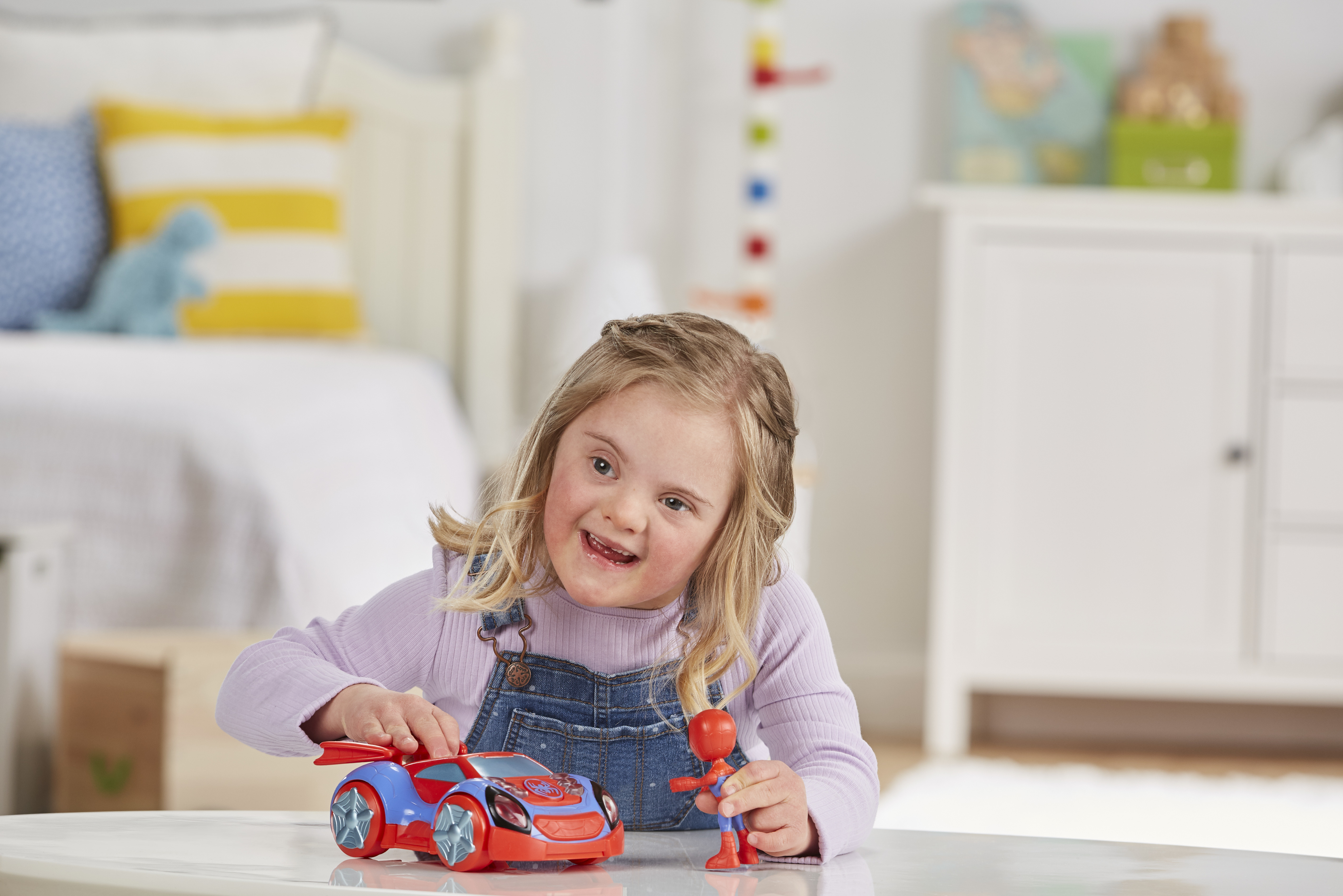 Dziewczynka uśmiecha się, trzymając zabawkowe auto i figurkę. W tle łóżko i komoda.