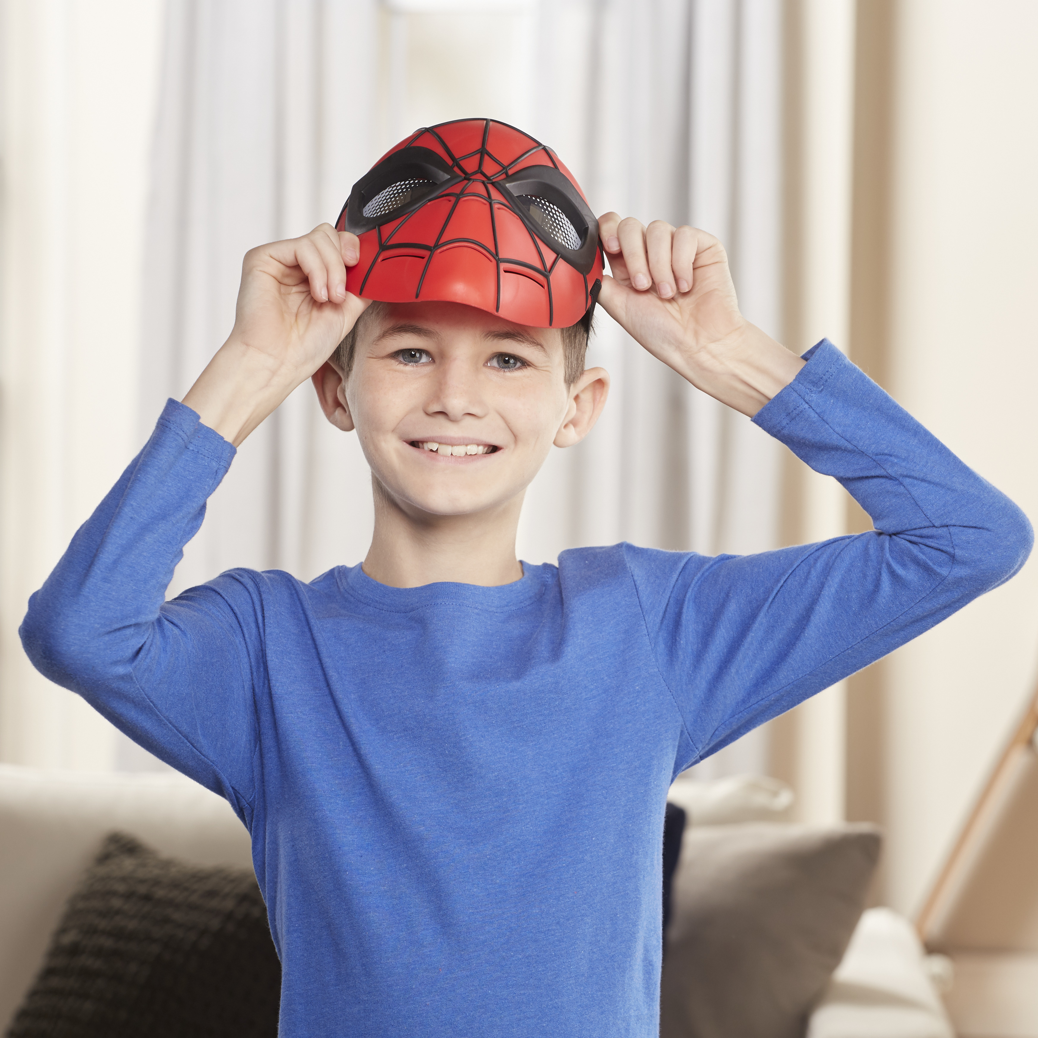 Ein Junge lächelt und hält eine Spiderman-Maske. Er trägt ein blaues Hemd, mit neutralem Hintergrund.