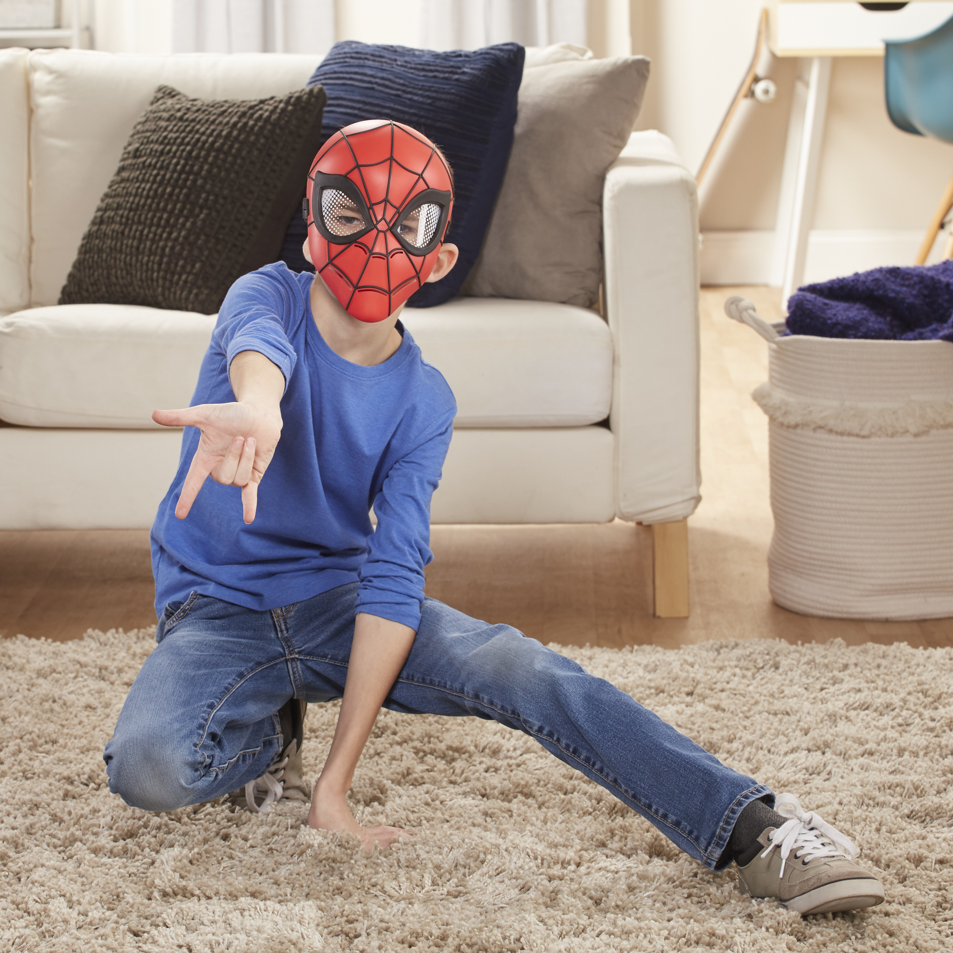 Ein Junge mit Spider-Man-Maske und blauem Hemd zeigt auf. Er kniet auf einem Teppich. Eine weiße Couch und ein Korb sind im Hintergrund.