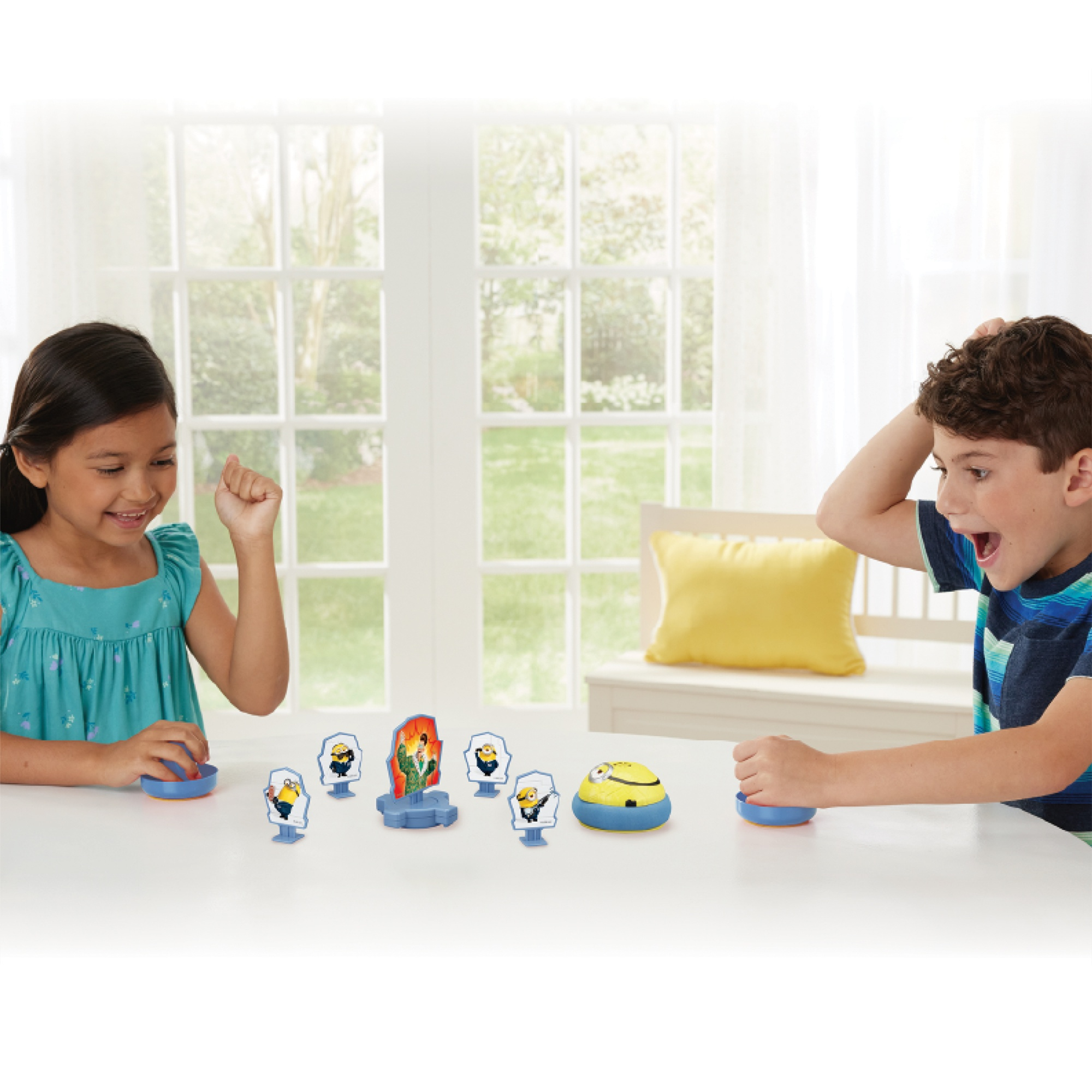 Due bambini giocano con figurine dei Minion e un dispositivo giallo su un tavolo bianco. Una finestra sullo sfondo.