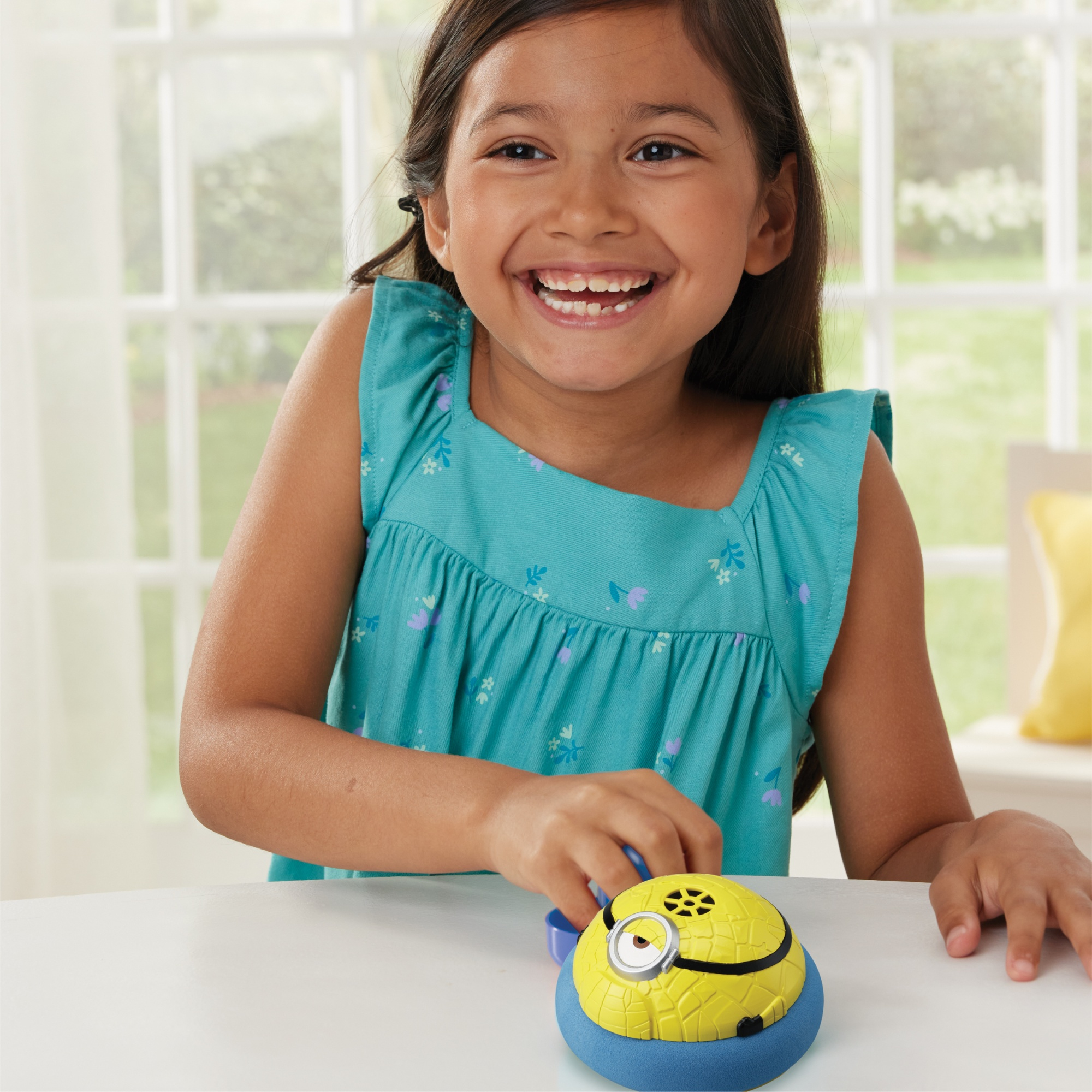 Una ragazza sorride, tenendo in mano un giocattolo Minion giallo su una base blu. Un tavolo bianco in primo piano, finestra e tende sullo sfondo.