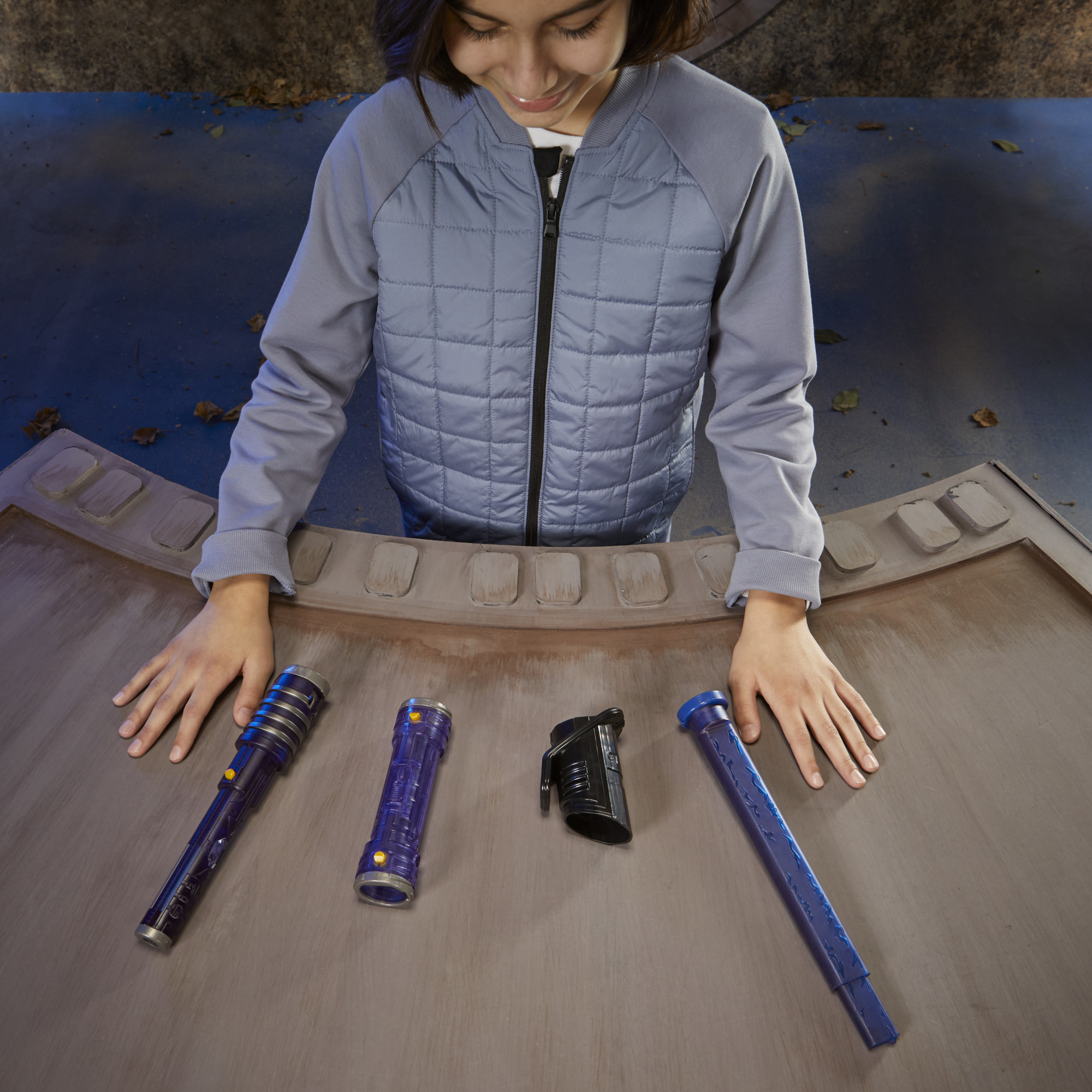 Osoba patrzy w dół na cztery zabawki miecze świetlne, niebieskie i czarne, na stole. Osoba nosi szarą kurtkę.