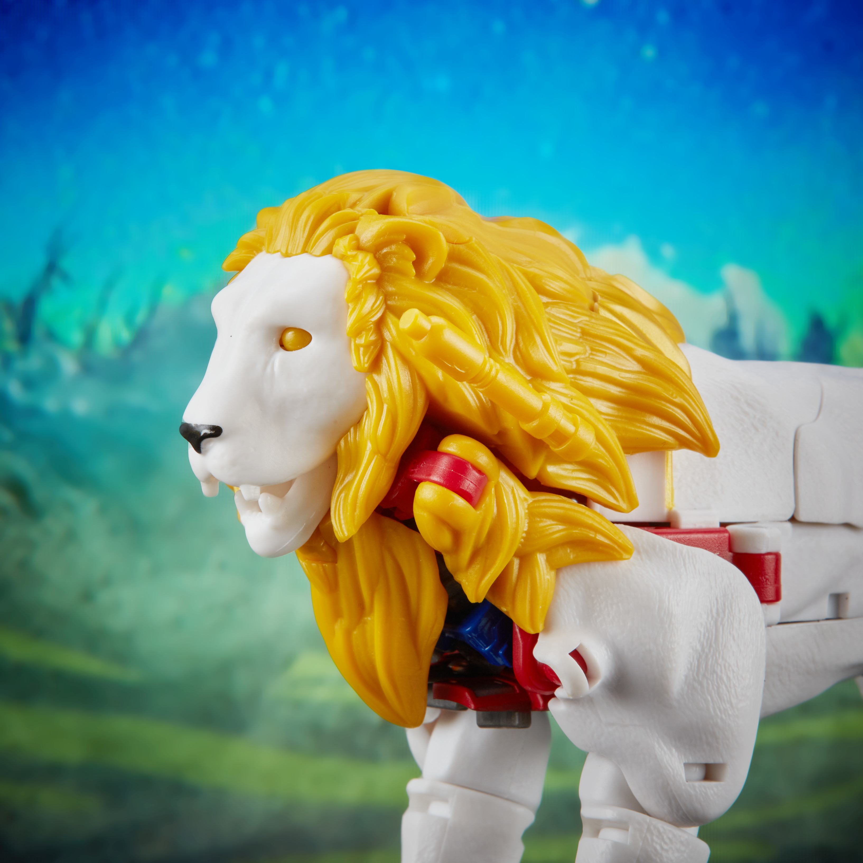 Biała zabawka Transformers w kształcie lwa z żółtą grzywą i czerwonymi detalami, na rozmytym niebiesko-zielonym tle.