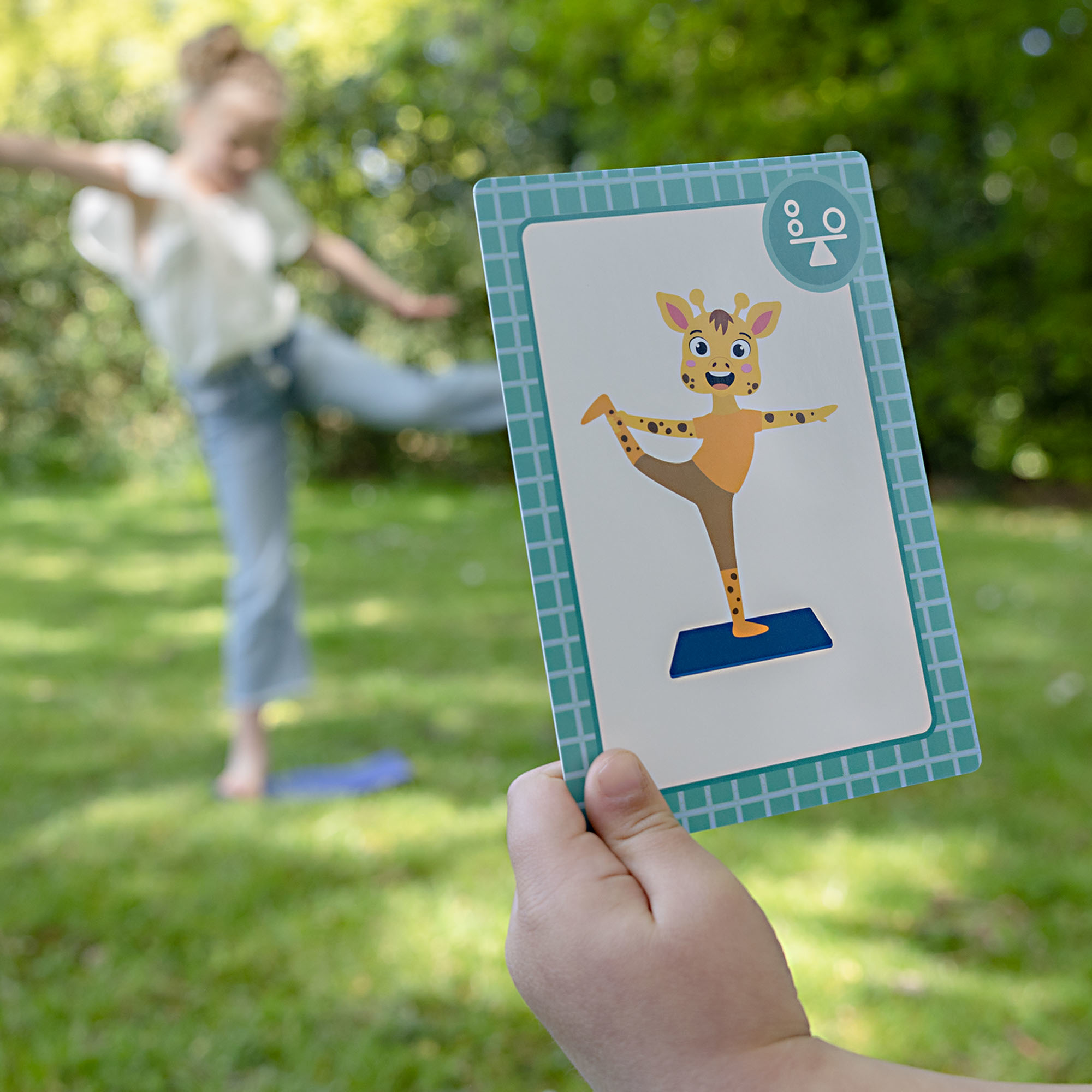 Viene mostrata una carta con una giraffa che fa yoga. Una ragazza si equilibra sullo sfondo. Sono visibili erba verde e alberi.