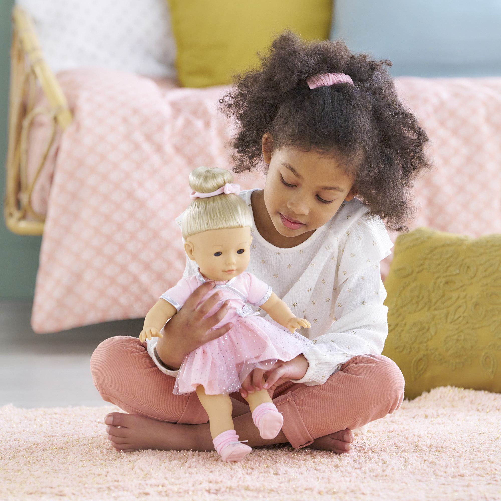 Ein Mädchen sitzt und hält eine Puppe. Die Puppe trägt ein rosa Kleid. Mädchen trägt weißes Hemd und rosa Hose. Rosa Teppich.