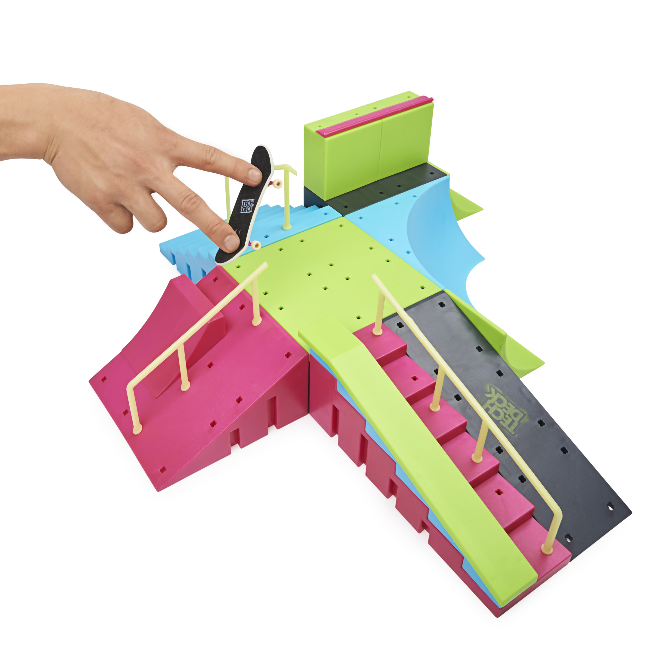 Una mano sostiene un monopatín en miniatura sobre un parque de patinaje modular y colorido con rampas, rieles y escaleras.