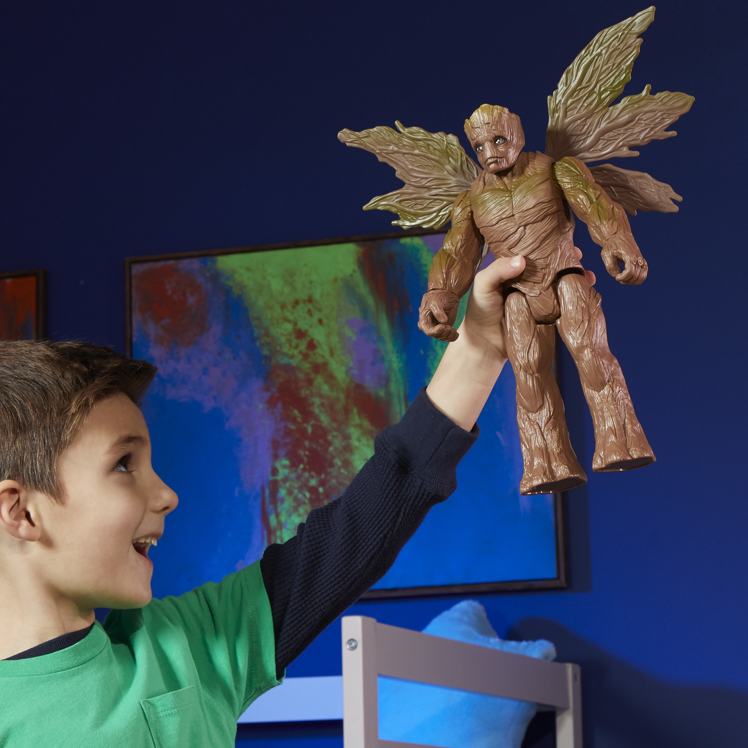 Un niño levanta un juguete de Groot con alas. El fondo tiene una pared azul y una pintura colorida.