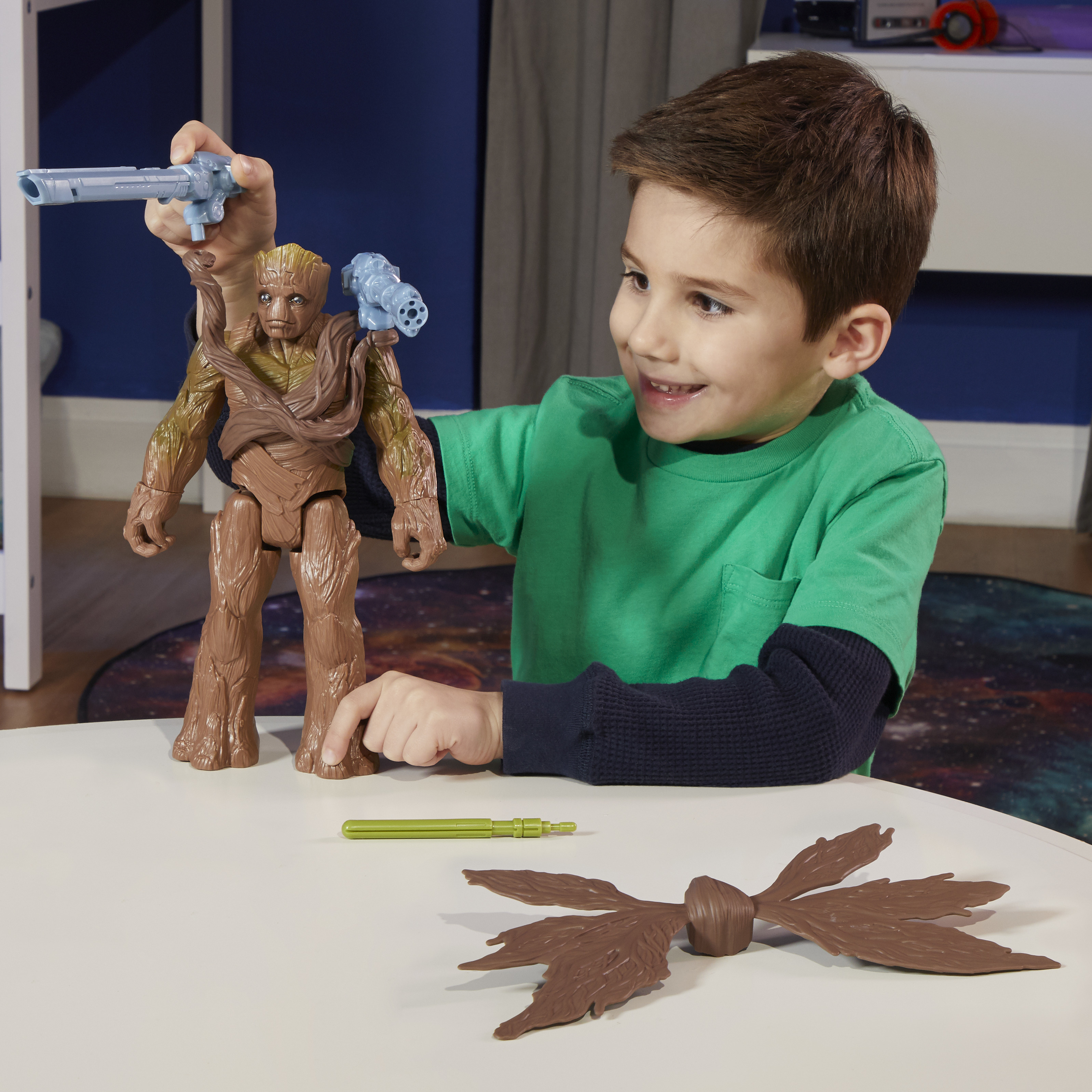 Un niño sonríe, sosteniendo un juguete de Groot con armas. Otras piezas del juguete están sobre la mesa. El fondo es una habitación.