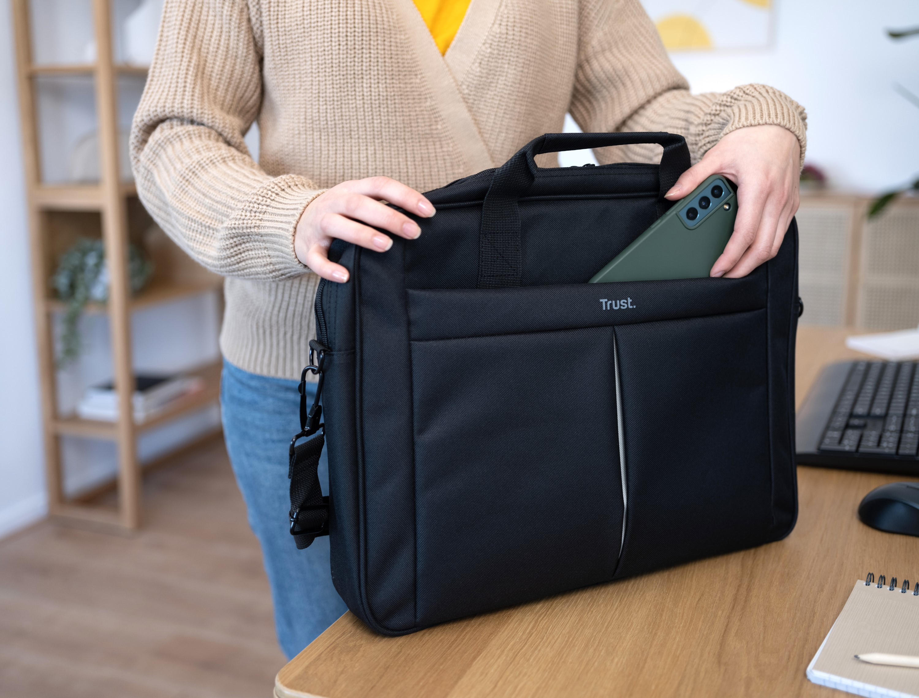 Una persona tiene una borsa nera per laptop con un telefono verde in tasca. Sulla borsa c'è la scritta 'Trust'.