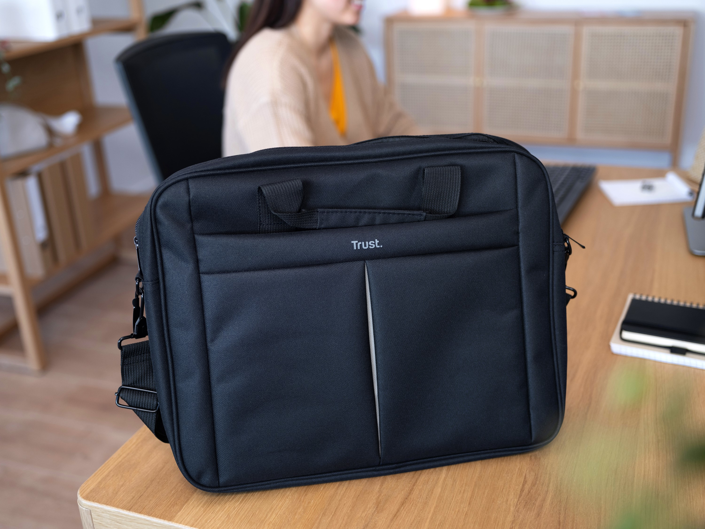 Borsa per laptop Trust nera su scrivania in legno. Una persona lavora sullo sfondo.