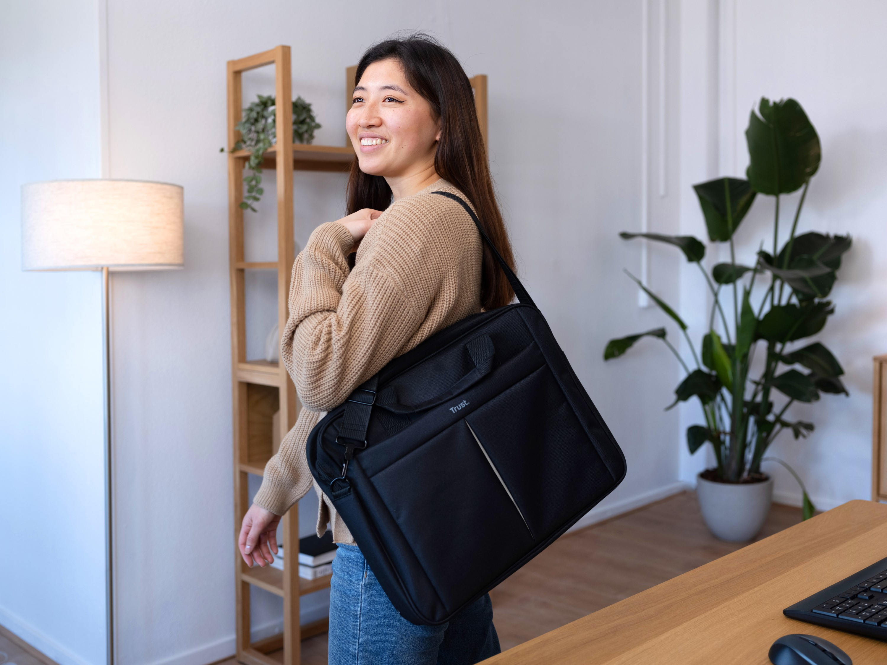 Una donna sorridente porta una borsa nera per laptop. È in una stanza con una mensola di legno e una pianta.