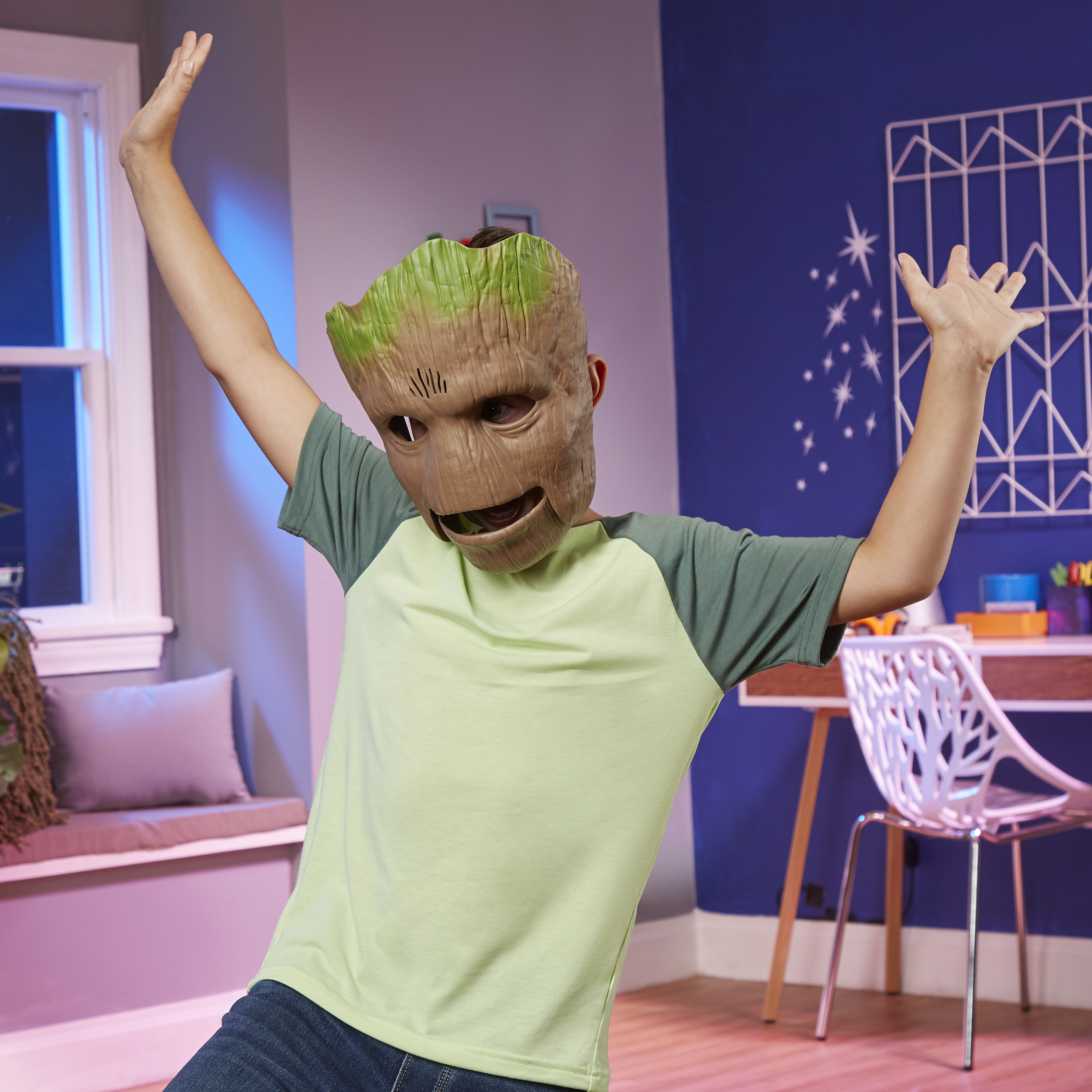 Una persona con una máscara de Groot levanta los brazos en una habitación. Un escritorio y una ventana están al fondo.