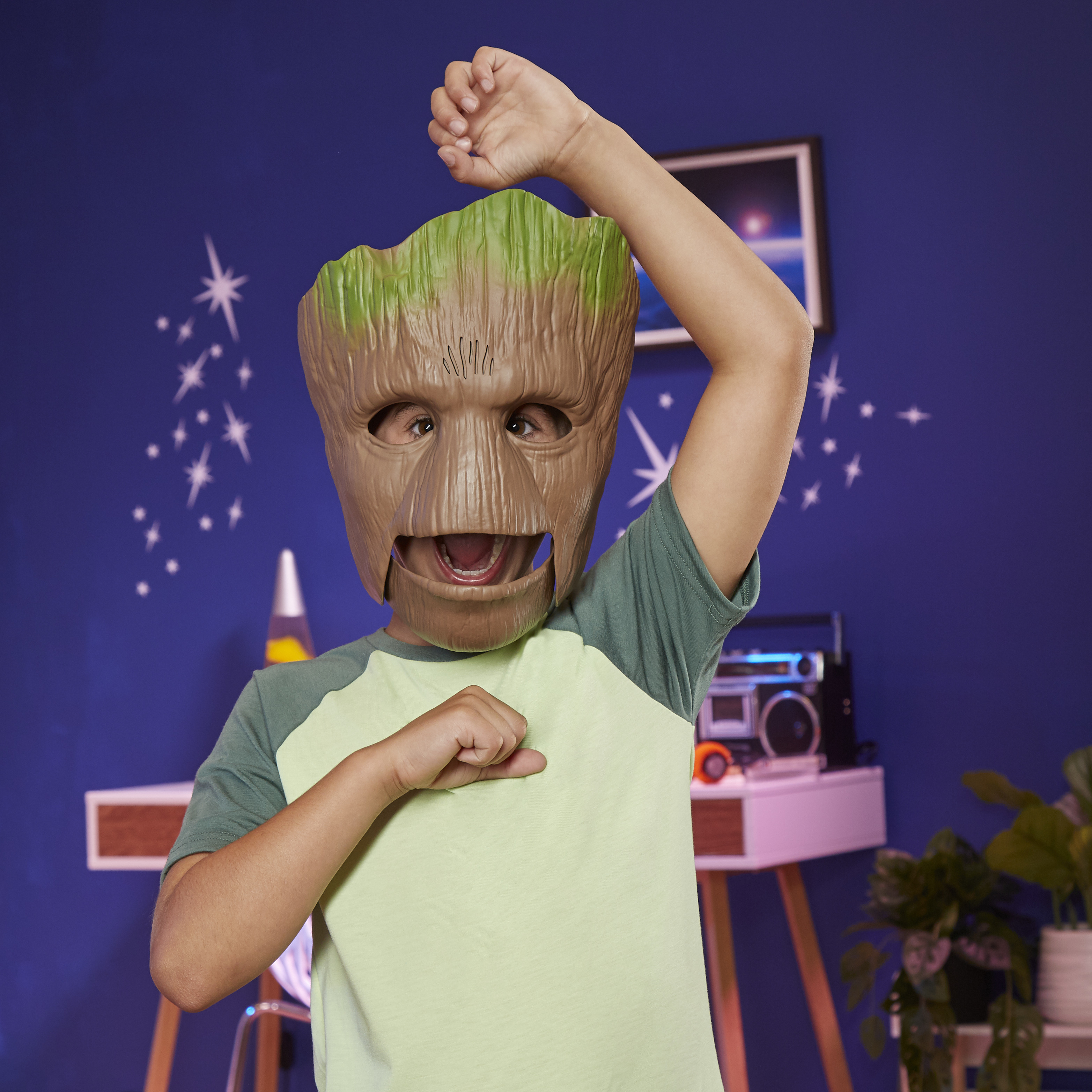 Un niño con una máscara de Groot levanta un brazo. El fondo muestra una pared azul con estrellas y una estantería.