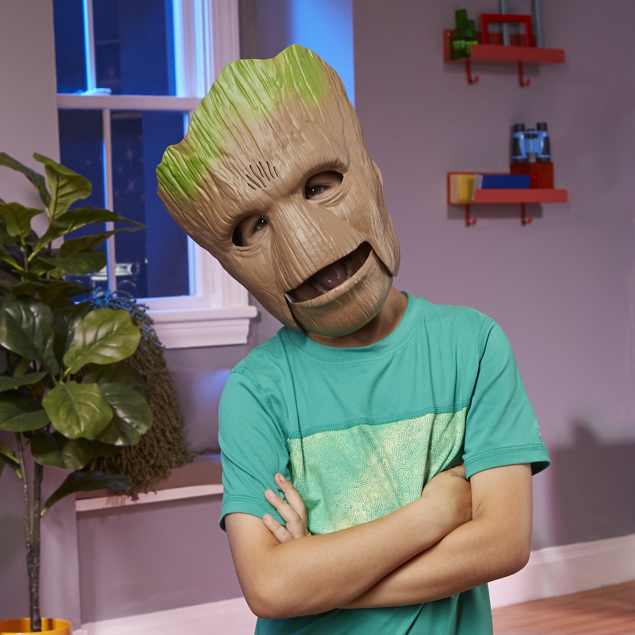 Un niño con una máscara de Groot y una camisa verde, con los brazos cruzados. El fondo incluye una ventana, estantes y una planta.