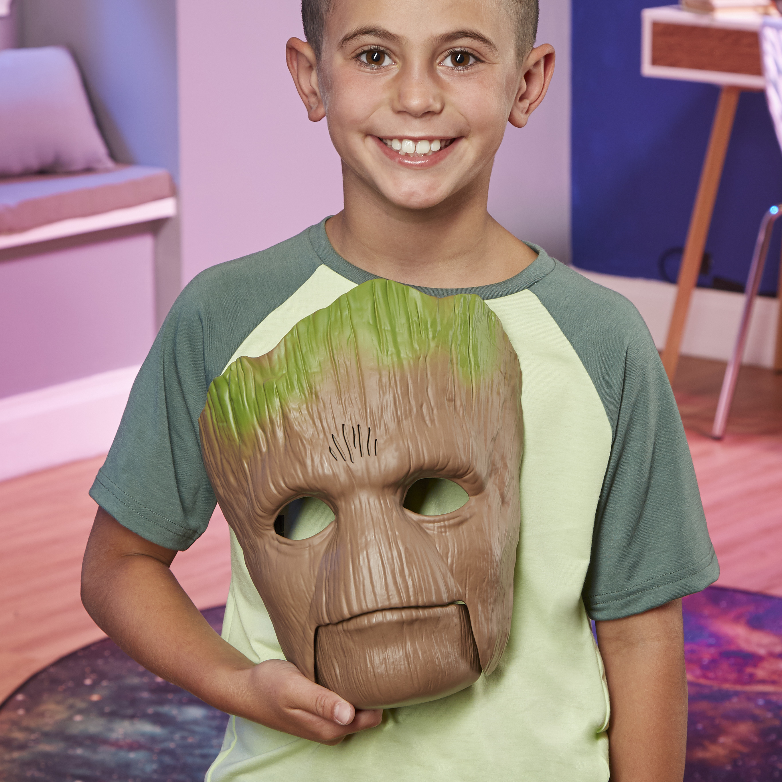 Un niño sonríe, sosteniendo una máscara de Groot. Lleva una camisa verde. El fondo tiene un escritorio y una pared azul.