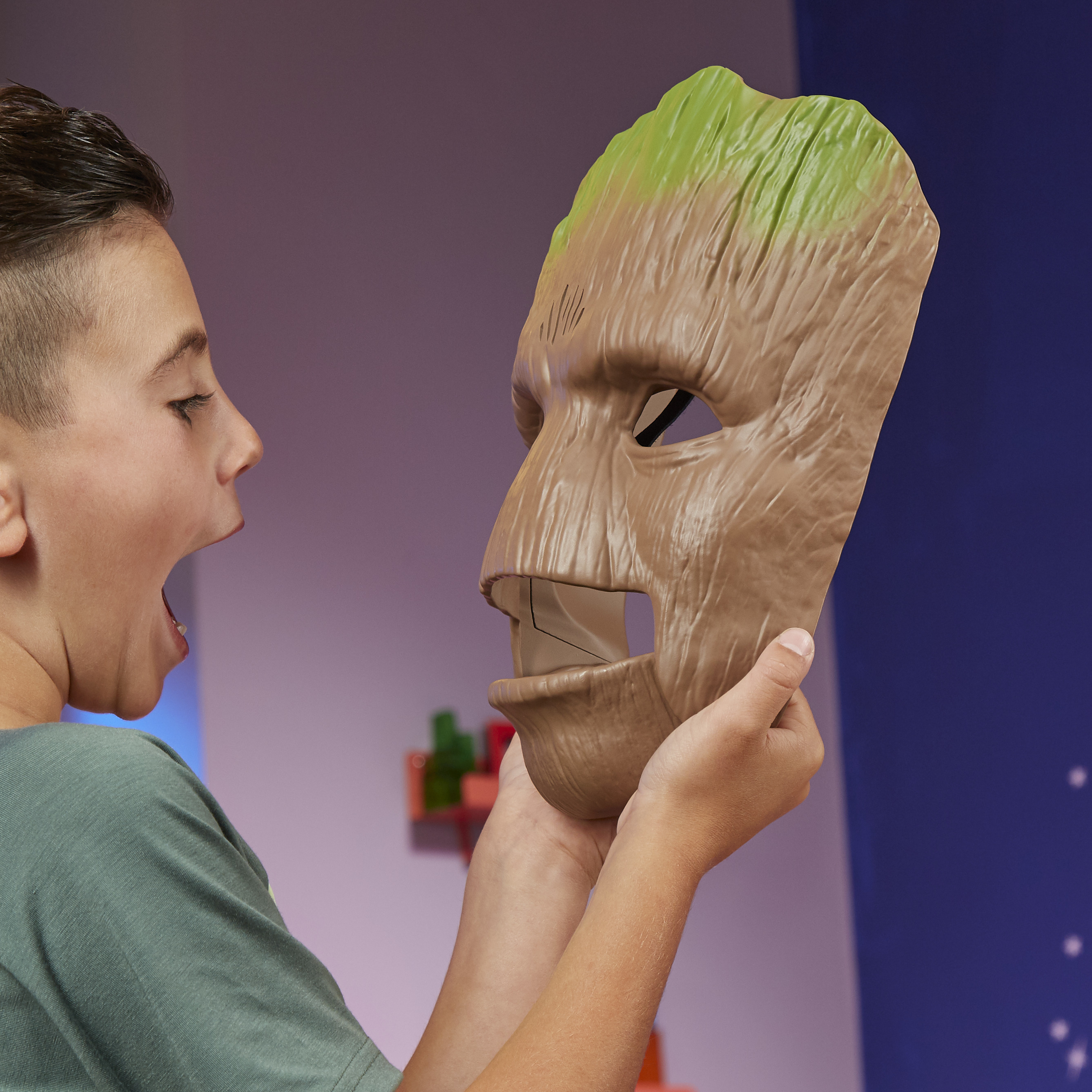 Un niño sostiene una máscara de Groot, mirándola con la boca abierta. La máscara es marrón y verde. El fondo es morado y azul.