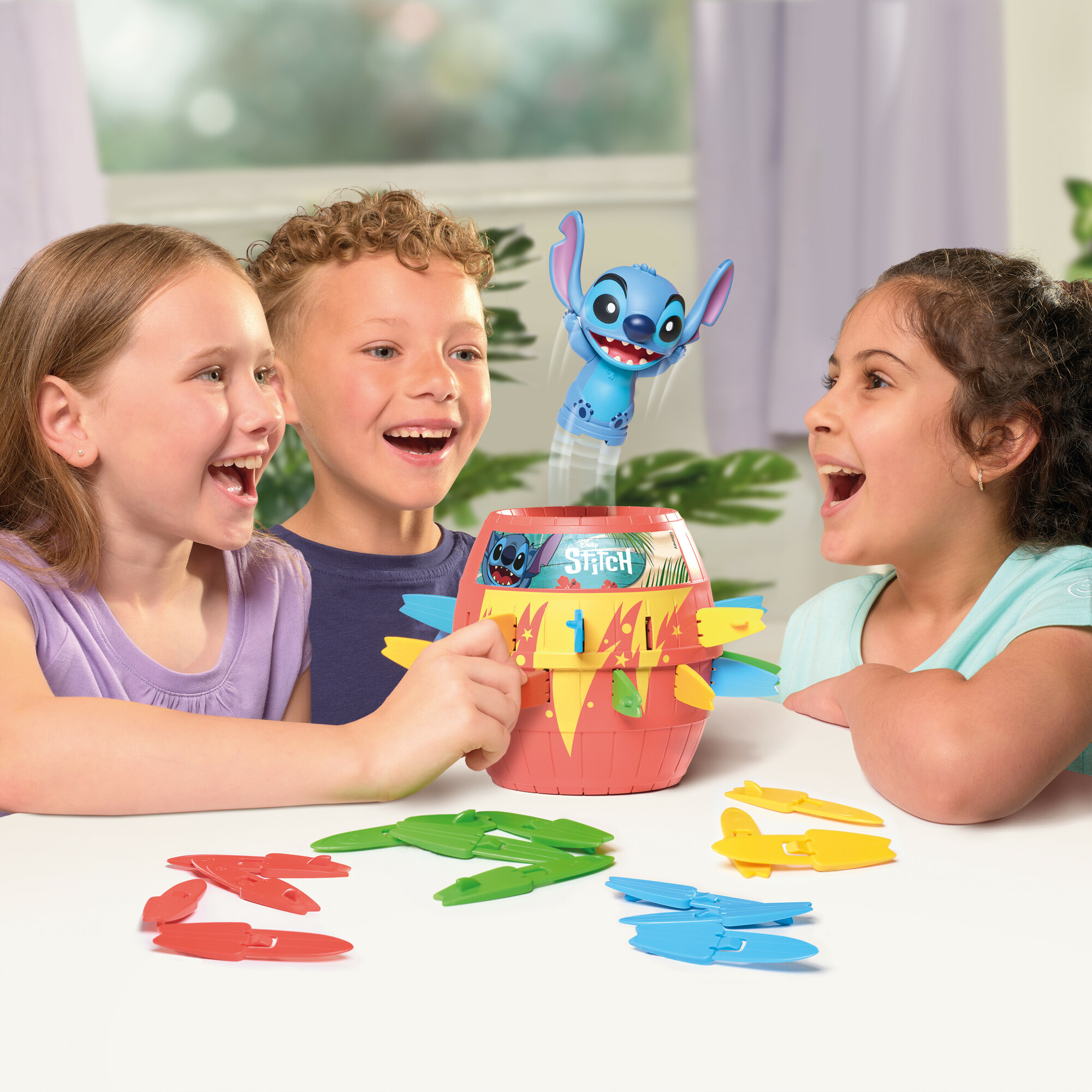 Trois enfants jouent à un jeu sur le thème de Stitch. Un jouet Stitch bleu saute d'un tonneau. Différentes pièces de jeu colorées sont sur la table.