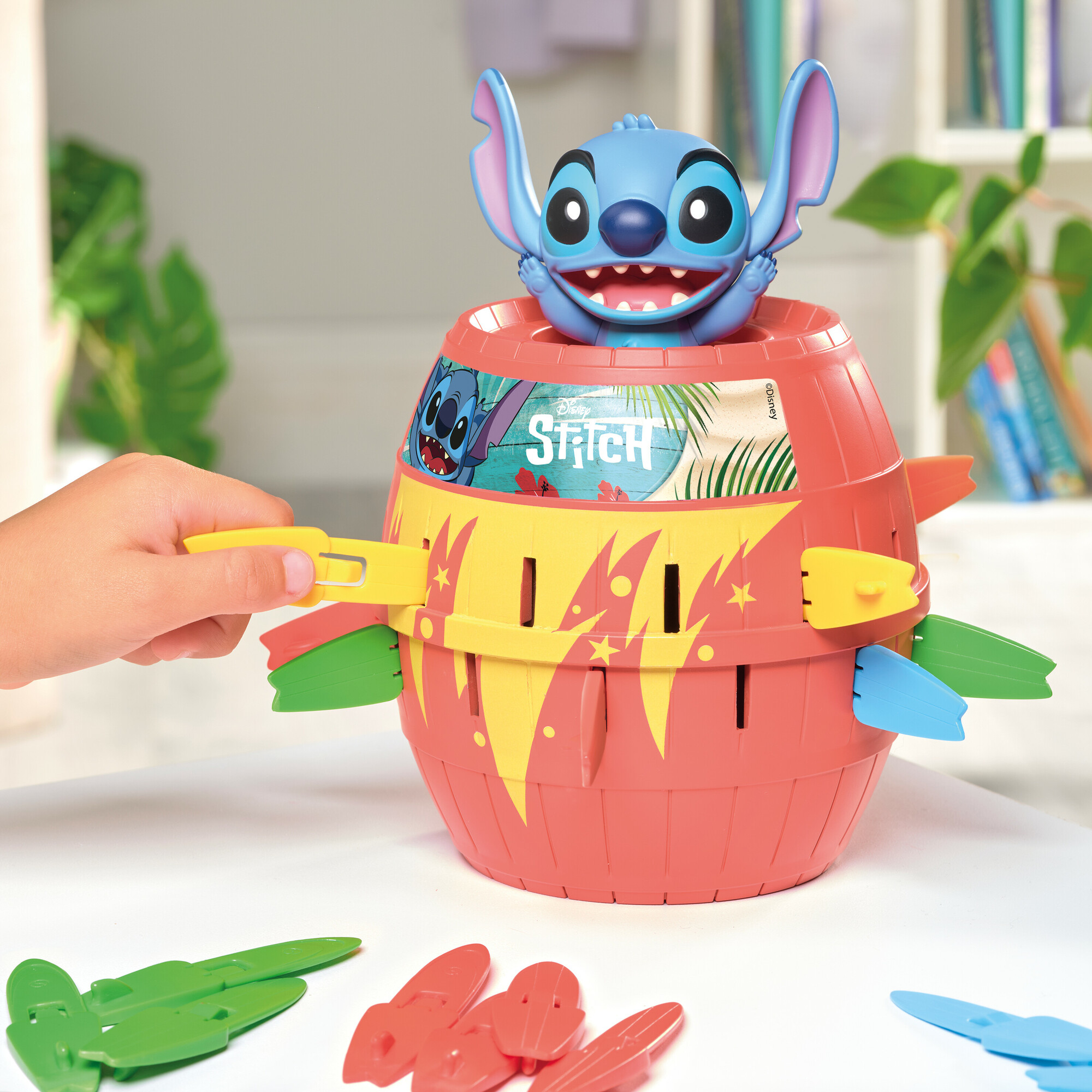 Un jeu sur le thème de Stitch avec une figurine bleue sur le dessus. Une main insère une pagaie jaune. Plusieurs pagaies colorées sont présentes.
