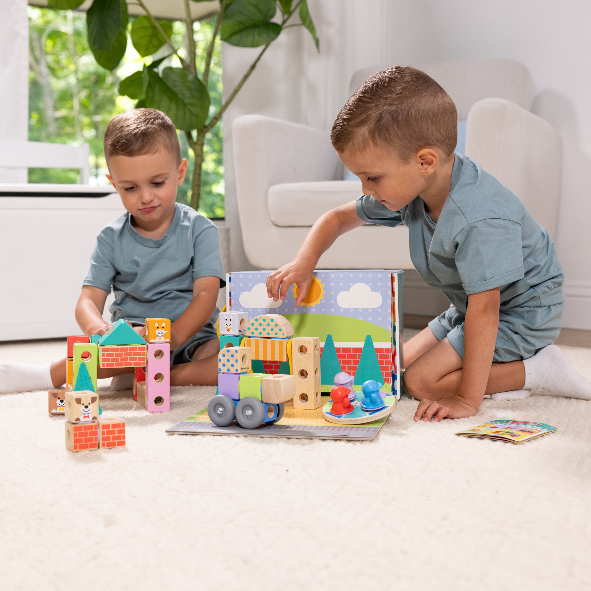 Due ragazzi giocano con blocchi di costruzione. Uno costruisce una casa, l'altro interagisce con una scena giocattolo. Su un tappeto.