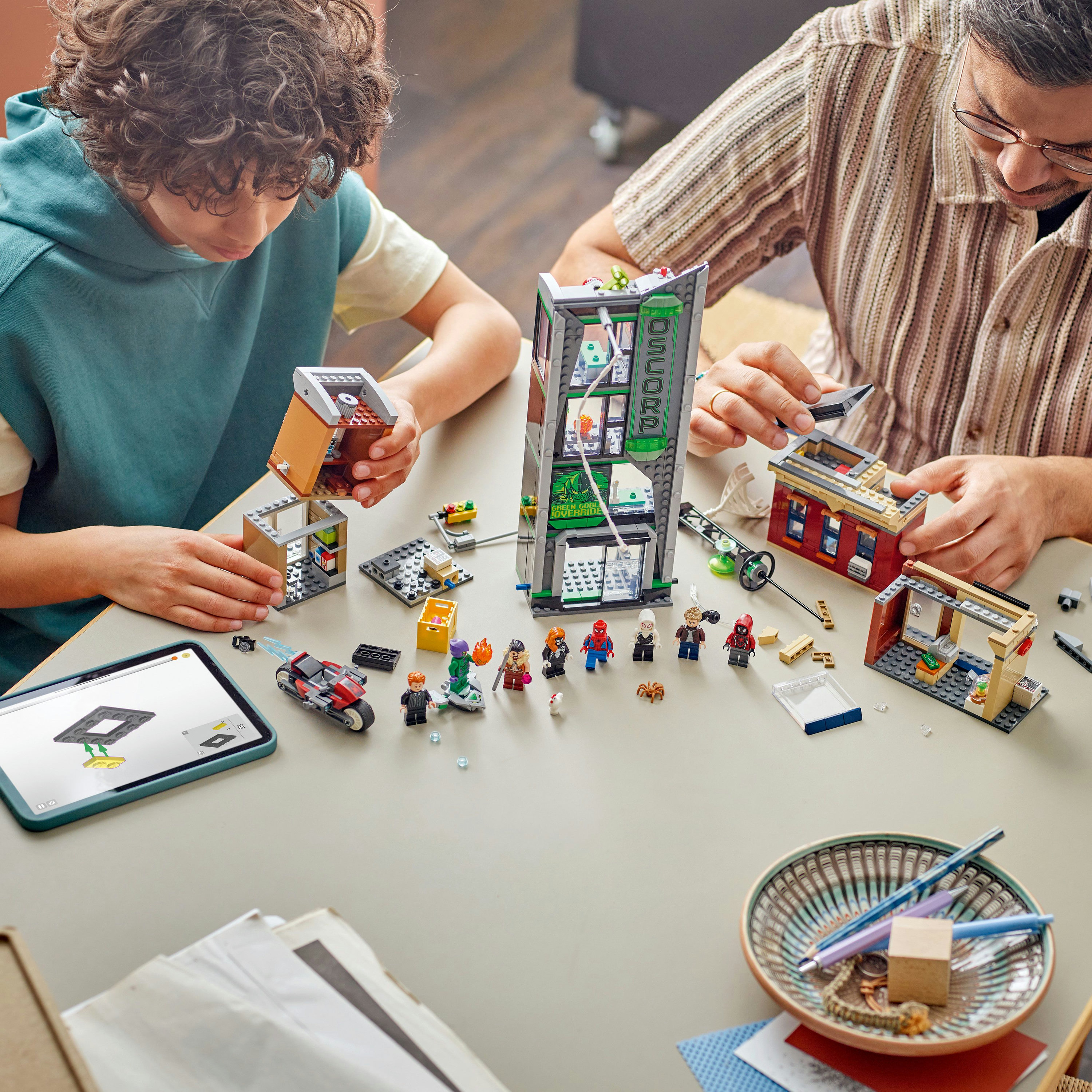 Dwie osoby budują zestaw Lego z tabletem pokazującym instrukcje, różne figurki i budynki Lego.
