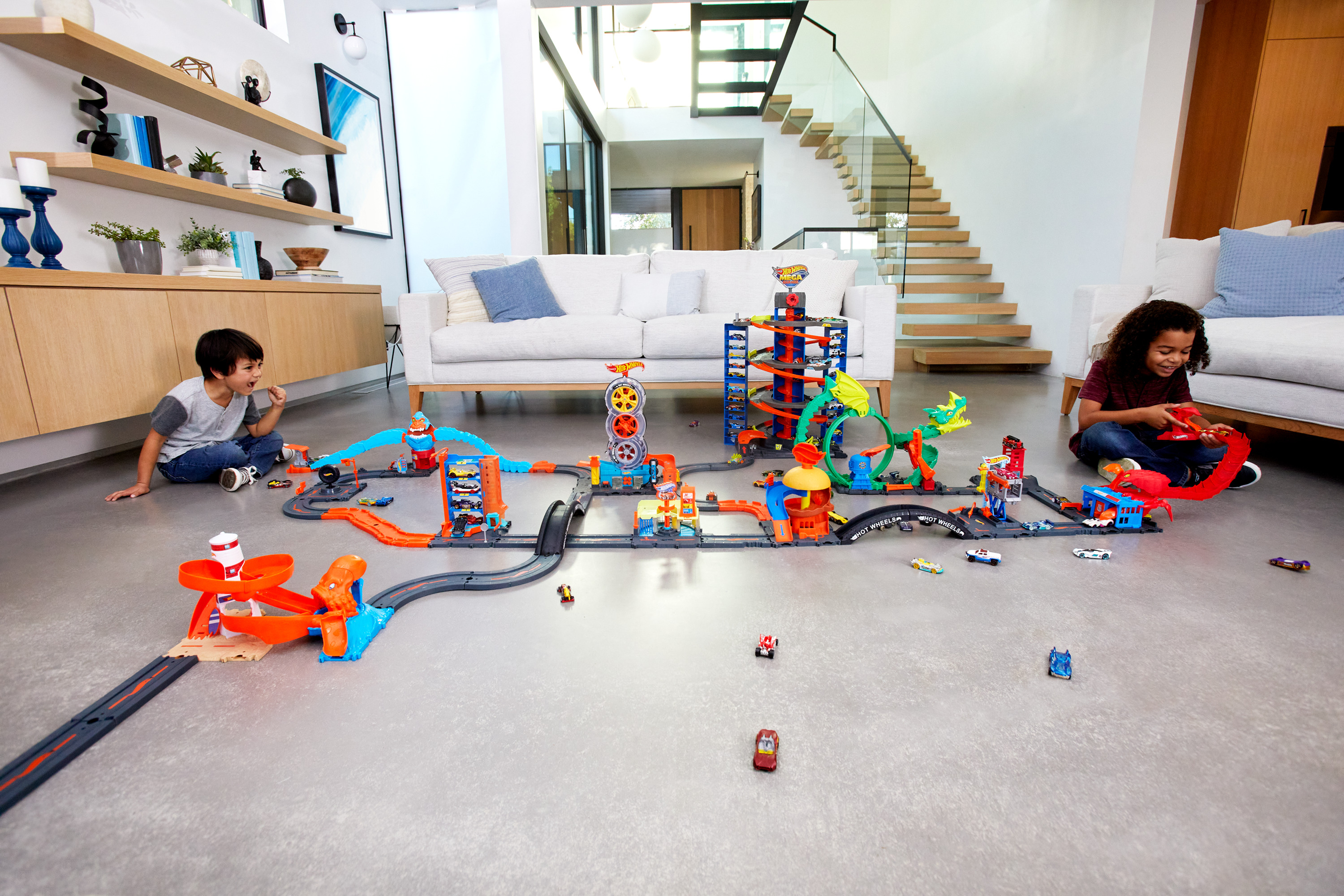 Dwoje dzieci bawi się zestawem torów Hot Wheels w nowoczesnym domu. Zestaw jest na podłodze i zawiera samochody i tory.
