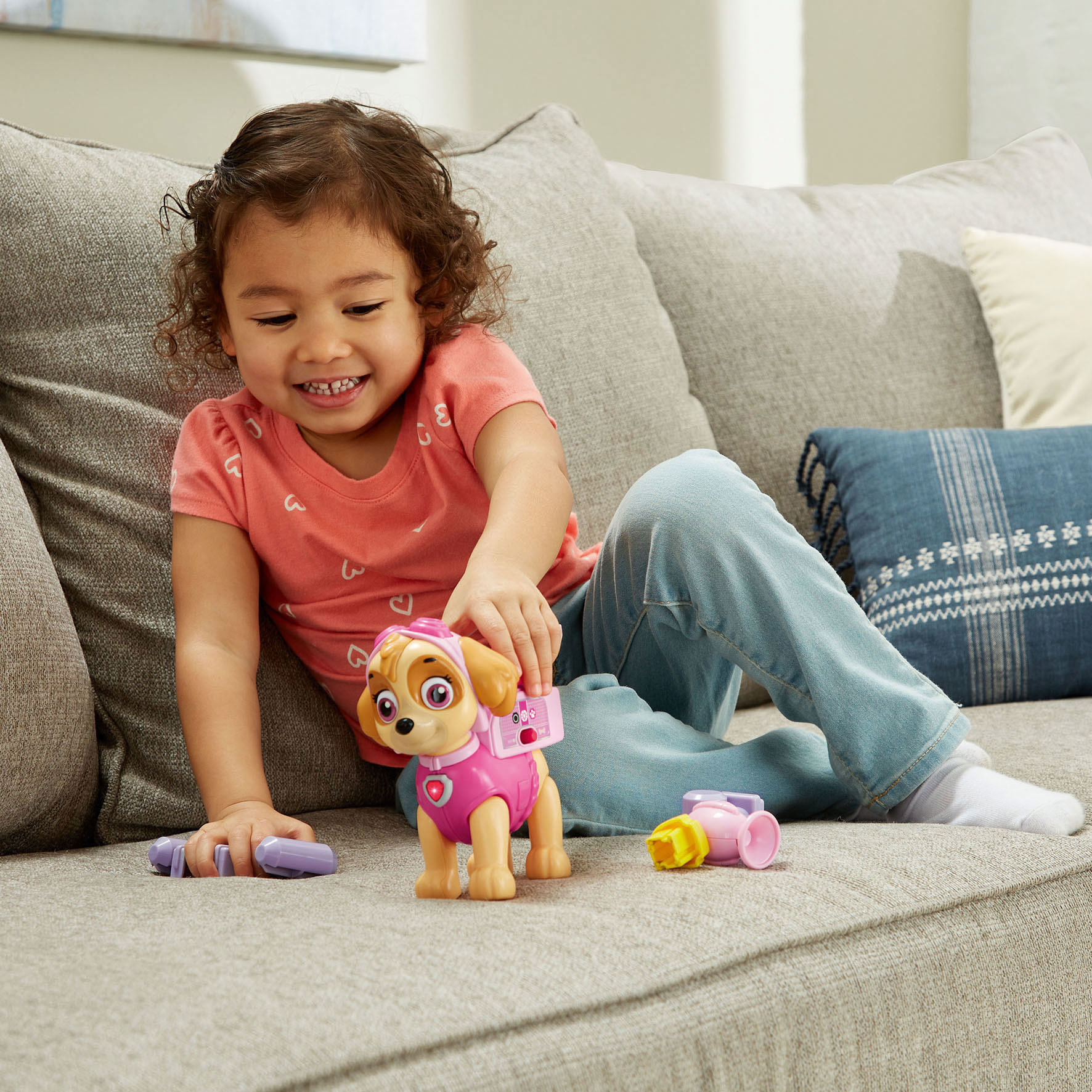 Ein lächelndes Kind spielt mit einem Skye-Spielzeug auf einem grauen Sofa. Spielzeug und Kissen sind ebenfalls vorhanden.