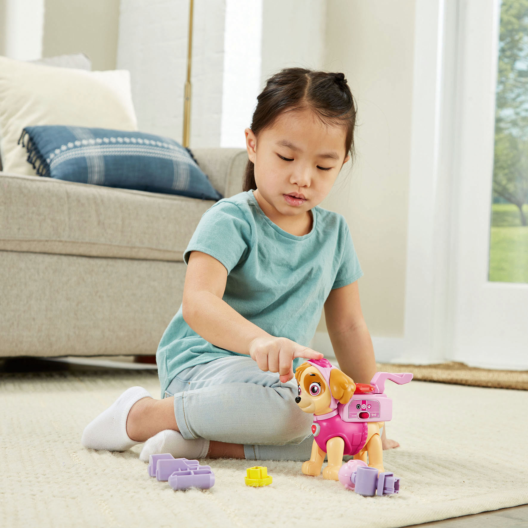 Ein Mädchen spielt mit einem Skye-Spielzeug von PAW Patrol. Sie sitzt auf einem Teppich, im Hintergrund eine Couch.