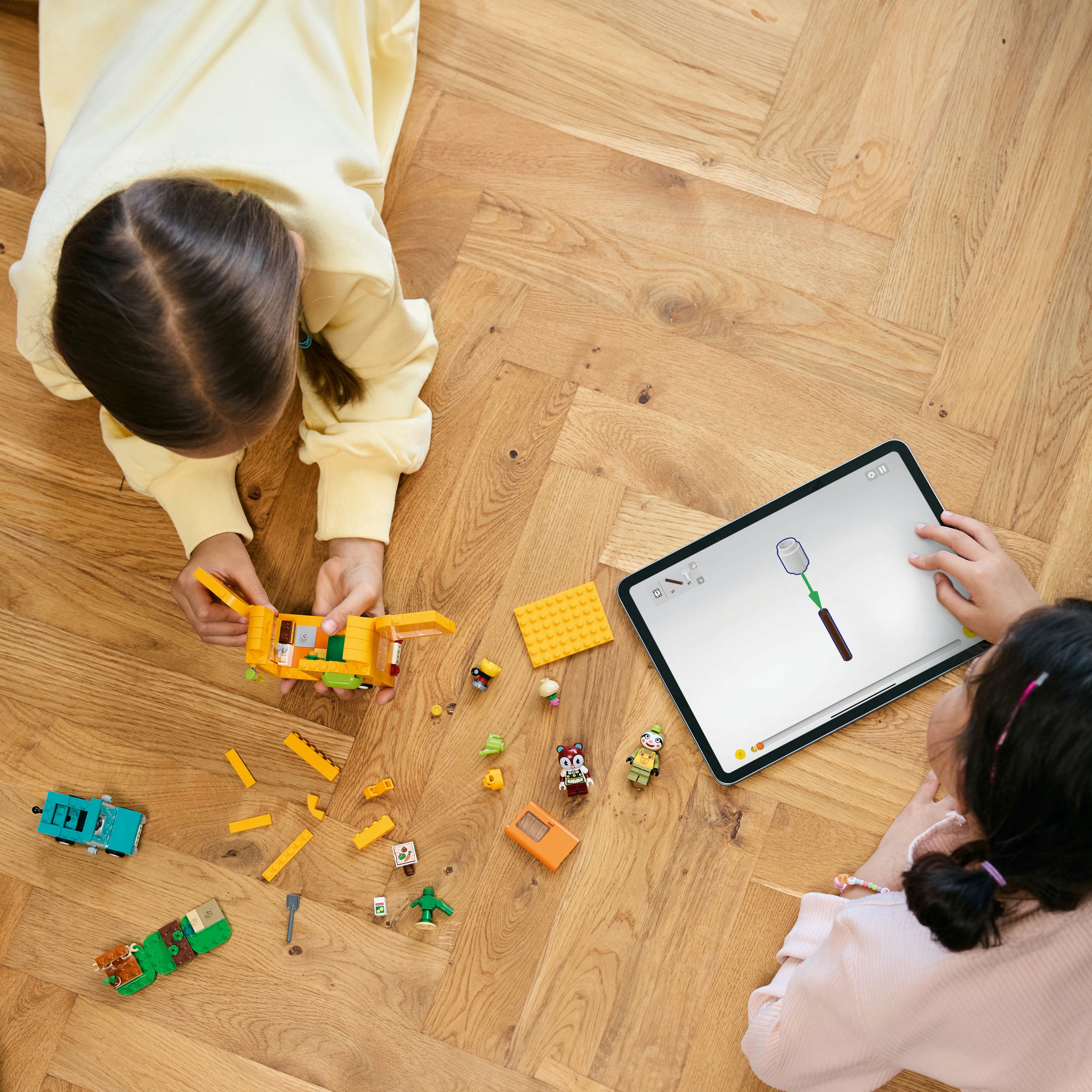 Dwoje dzieci na drewnianej podłodze bawią się Lego i tabletem. Klocki i figurki Lego są rozrzucone.