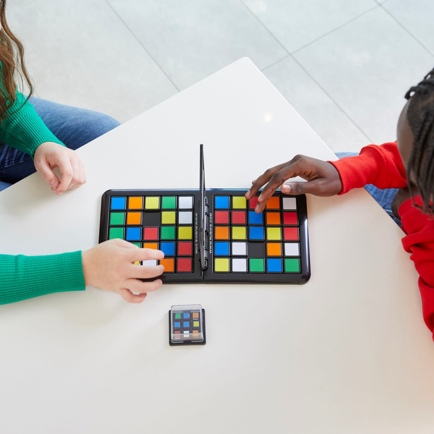Dos personas juegan un juego de mesa con cuadrados coloridos sobre una mesa blanca. Un cubo pequeño está debajo. Uno viste de verde, el otro de rojo.