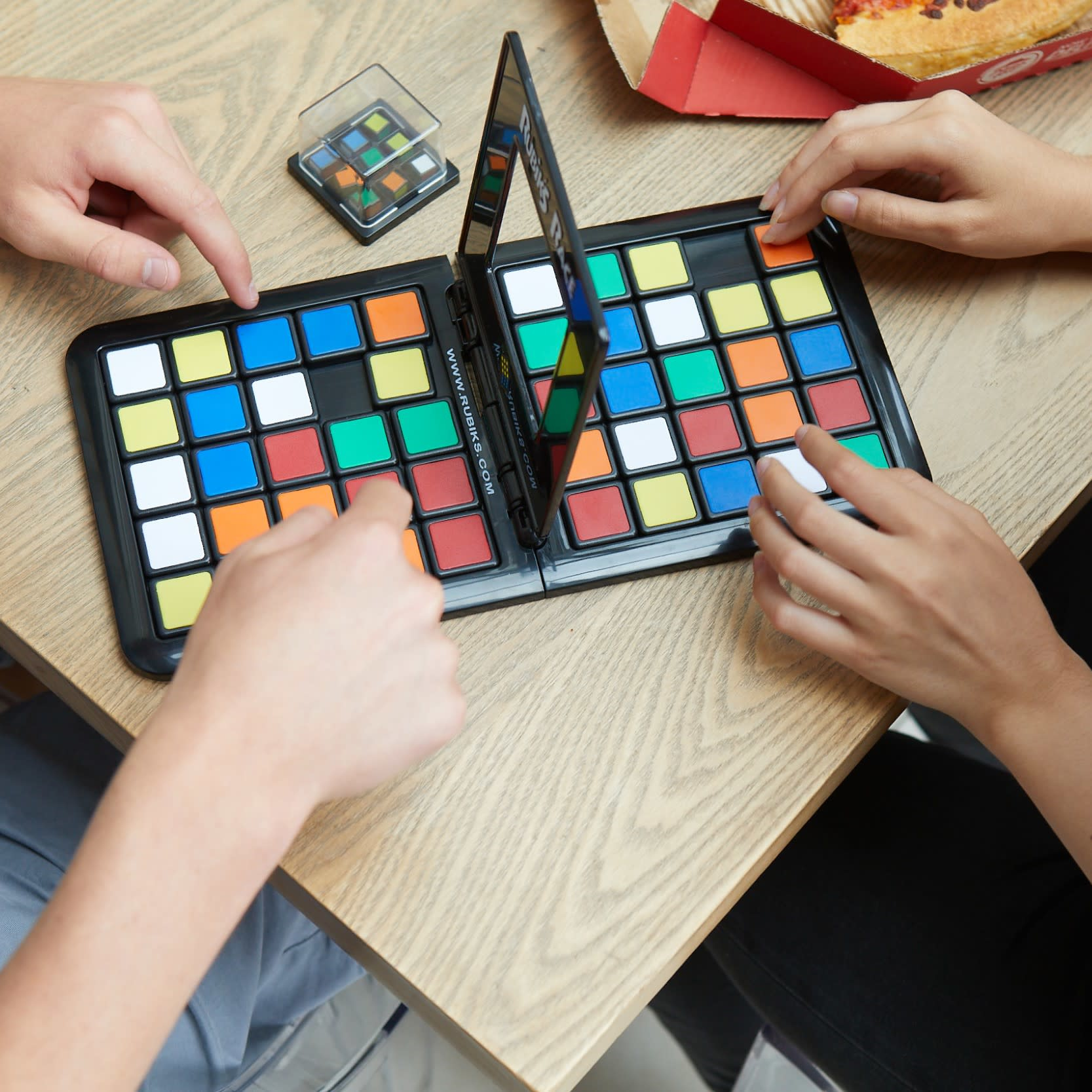 Personas jugando Rubik's Race con cuadrados de colores en una mesa de madera, caja de pizza al fondo.