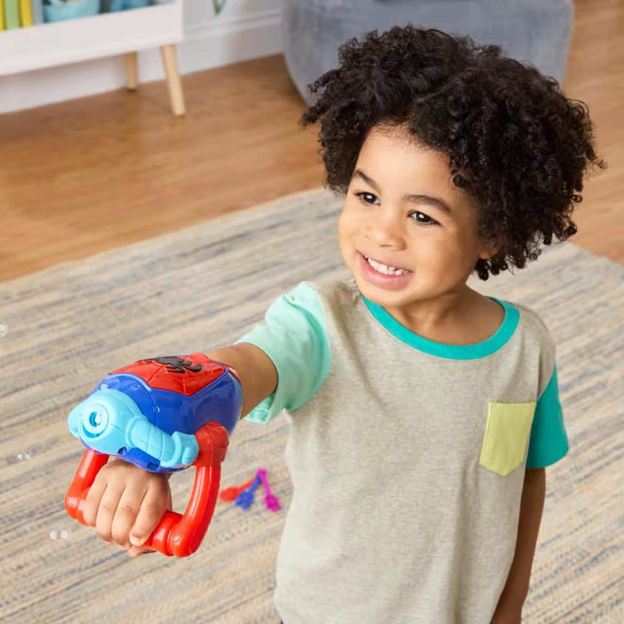Un niño sonriente sostiene una pistola de juguete. La pistola es roja, azul y blanca. El niño lleva una camisa gris.