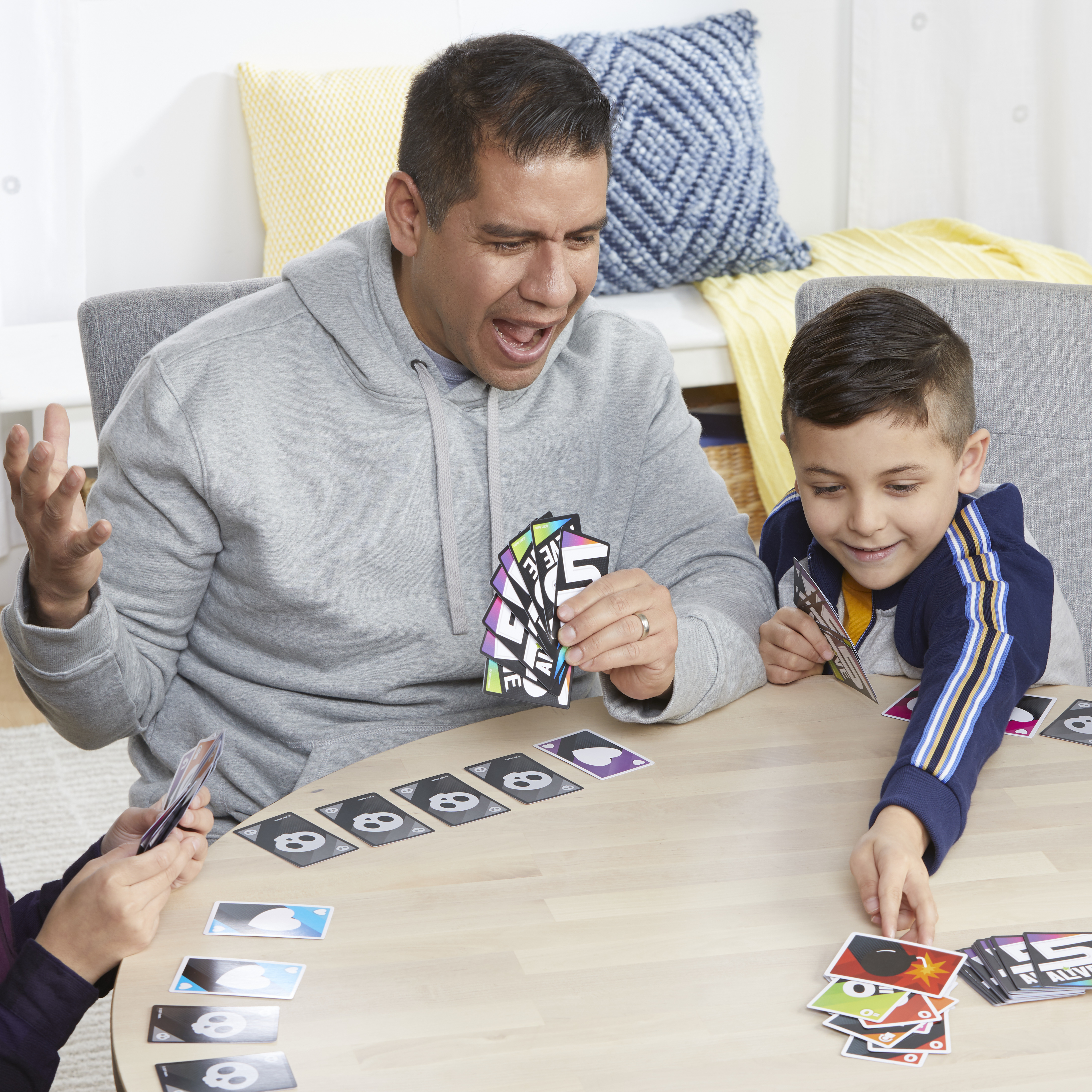 Drei Personen spielen ein Kartenspiel. Ein Mann und ein Junge halten Karten. Karten mit Totenköpfen und anderen Bildern liegen auf dem Tisch.