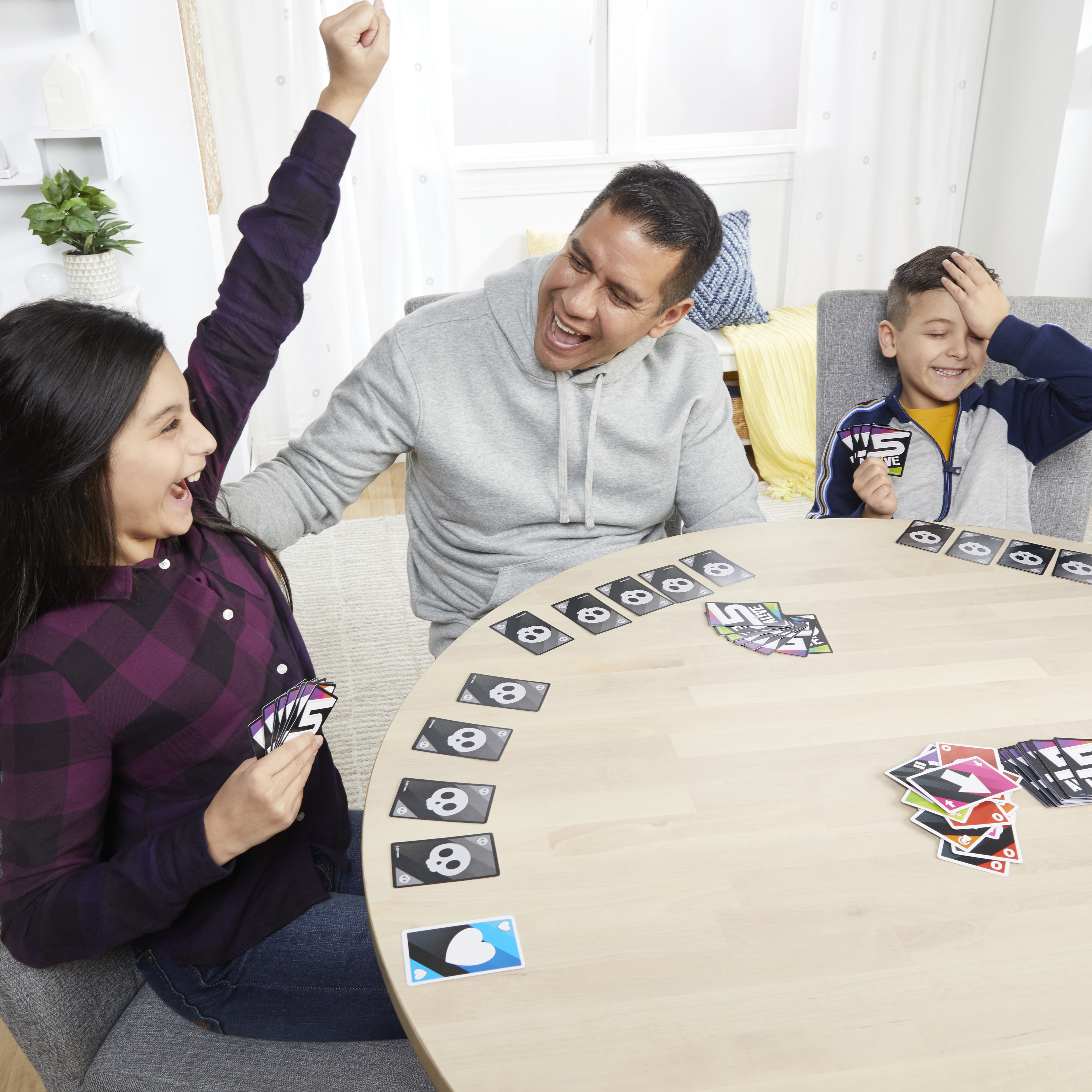 Familie spielt Kartenspiel. Mädchen hebt Arm, Vater lacht, Junge bedeckt Gesicht, alle am Tisch.