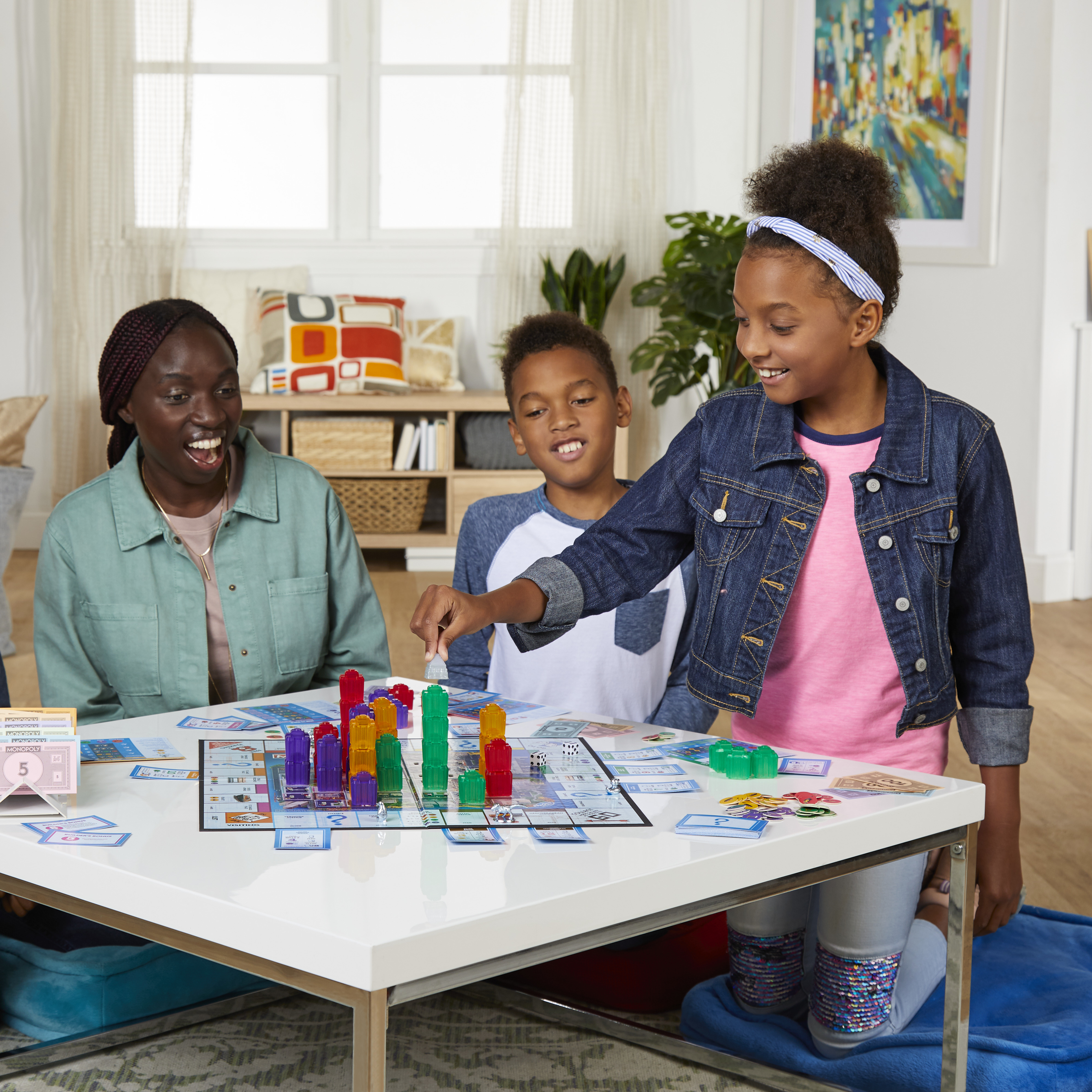 Drie kinderen en een volwassene spelen Monopoly. Het meisje in spijkerbroek wijst naar het bord. Kleurrijke gebouwen staan erop.
